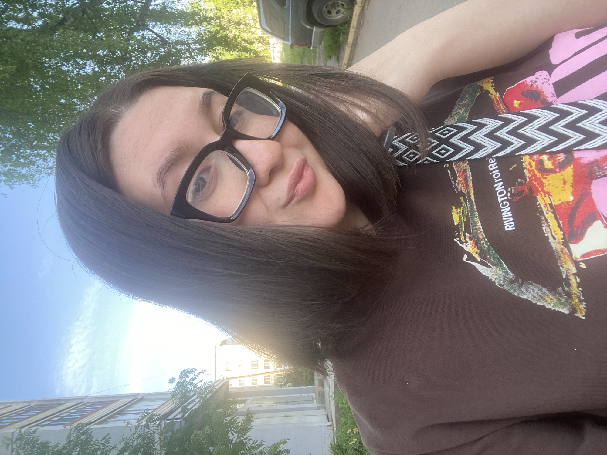 A woman with dark hair and black glasses is taking a selfie outdoors. She is wearing a brown shirt with a colorful graphic design and a black and white patterned strap. The background includes a building with windows and trees, and a vehicle is visible in the distance. The image appears to be taken in Chaikovsky, Russia, based on the surrounding architecture and vegetation. The sky is light blue and sunny.