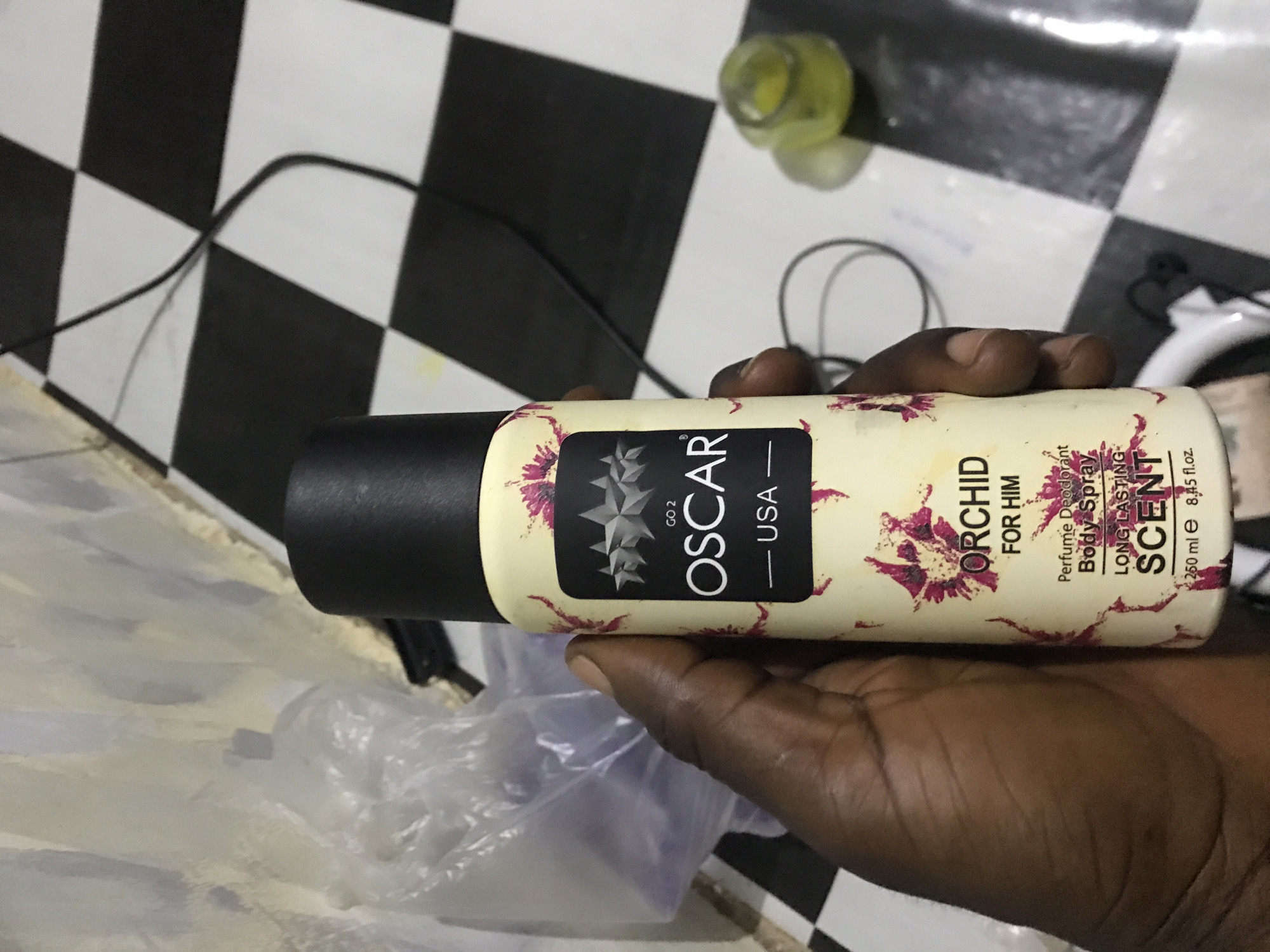 A dark-skinned right hand holds a cream-colored aerosol can with a black cap. The can displays a black rectangular label featuring the text "GO 2 OSCAR USA" in white, accompanied by three graduated gray stars. The can's body is patterned with scattered magenta-pink floral or abstract splatters. Below the main label, additional text reads "ORCHID FOR HIM," "Perfume Deodorant Body Spray," "LONG LASTING SCENT," and "250 ml e 8.45 fl.oz."
The background on the upper left consists of a light-colored, textured wall. Adjacent to the wall, a crumpled, translucent plastic bag containing indistinct items is visible. The floor throughout the scene is covered with black and white checkered tiles. Several black electrical wires are present on the tiled floor, some appearing loosely coiled. In the lower right portion of the image, a small, clear glass container holds a yellow substance and casts a shadow on the tile. The scene is an indoor setting in Rafin Par, Nigeria.
