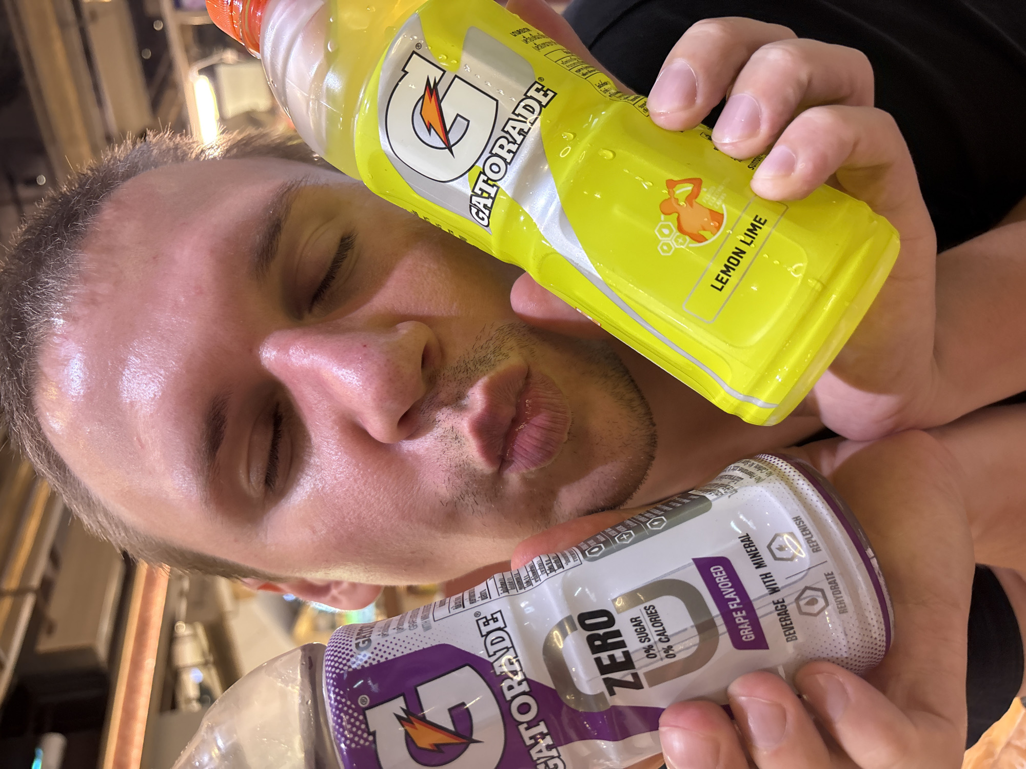 A close-up, overhead shot shows a person with their eyes closed and lips pursed, as if about to drink. The individual is holding two Gatorade bottles. One is a yellow "Lemon Lime" flavor, and the other is a purple "Grape Flavored" Zero Sugar variety. The bottles appear slightly damp, suggesting they may be cold. The setting seems to be indoors, possibly a store or gym, with blurred background elements that hint at shelving or equipment. The lighting is bright, and the overall mood is playful and casual. Visible text on the bottles includes "GATORADE," "LEMON LIME," "ZERO SUGAR," "0% CALORIES," and "GRAPE FLAVORED BEVERAGE WITH MINERAL." The presence of "SUNTORY" on the yellow bottle suggests a possible distributor or bottler. The image has a timestamp or watermark that appears to be from Chiang Mai City Municipality, Thailand.