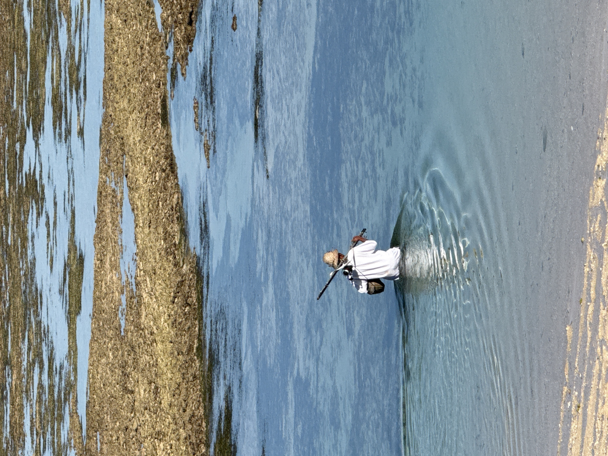A man in a white robe, tan hat, and carrying a rifle is wading through shallow water. The location is Kuta Selatan, Indonesia. The water ripples around him. To his left are strips of land; one sandy, one of brown seaweed or algae.