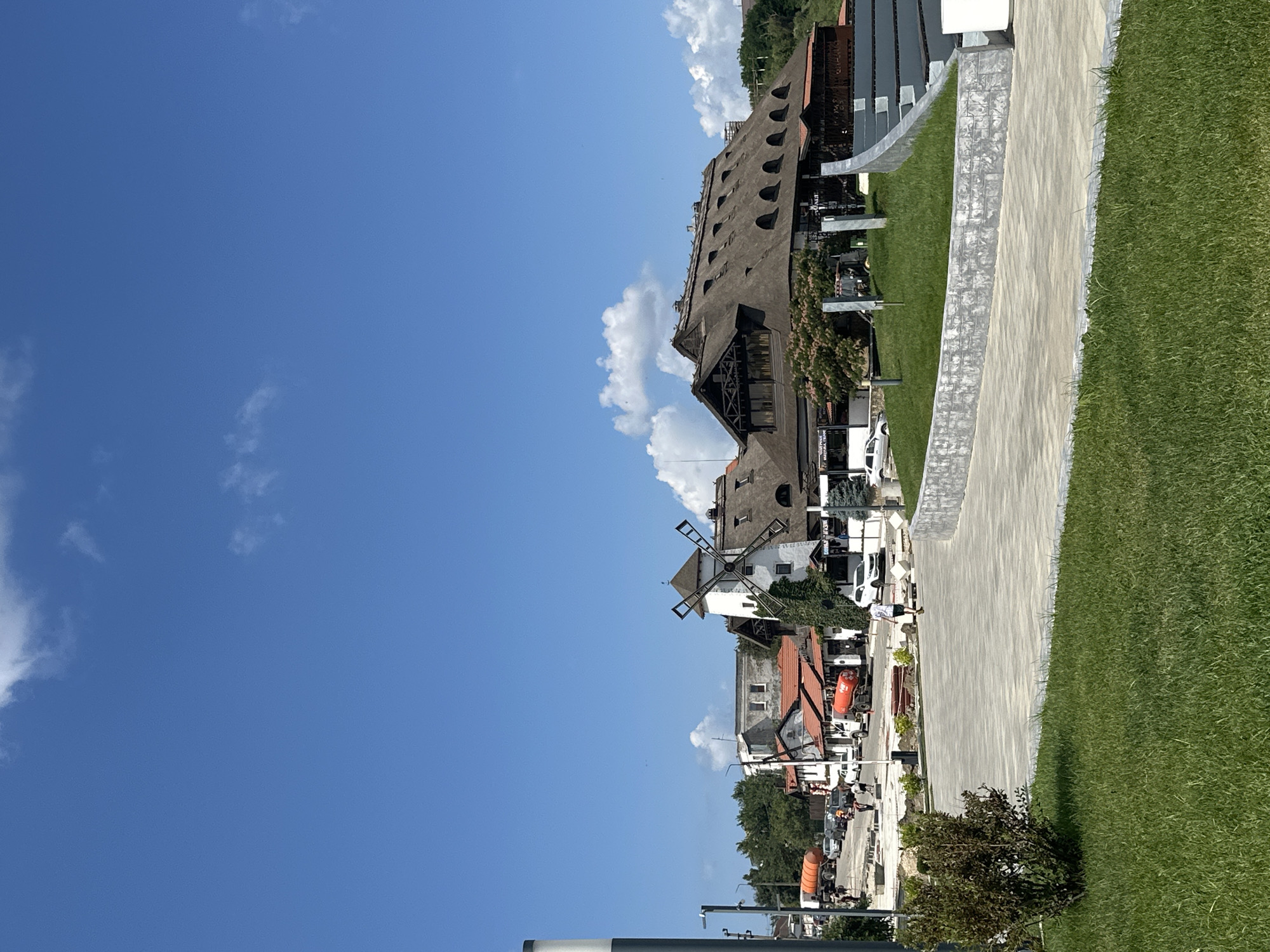 The image shows a bright, sunny day in Taman, Russia, with a clear blue sky and scattered white clouds. In the foreground, a manicured lawn leads to a paved walkway. Beyond the walkway, a street is visible with several buildings. A prominent structure is a white windmill with a dark thatched roof and visible wooden blades. Adjacent to the windmill is a large building with a prominent thatched roof that extends over a wide area, featuring numerous arched windows. Several cars are parked on the street, and two orange liquid tanker trucks are present. A few individuals are walking on the street and sidewalk. The scene appears to be a commercial or recreational area, possibly a tourist destination, given the unique architecture.