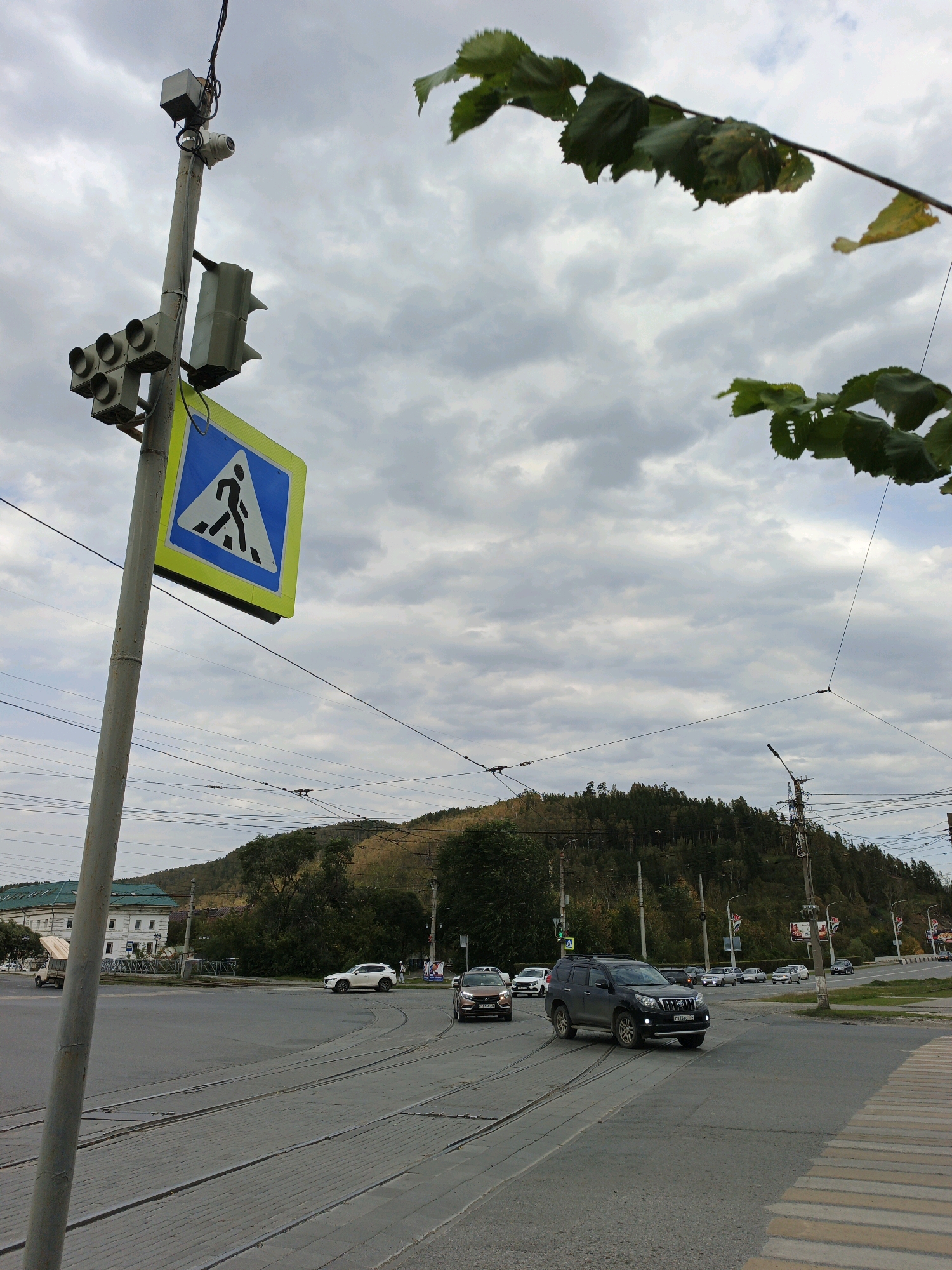 The image shows a street scene in Zlatoust, Russia, with a focus on a traffic light pole and an intersection. The traffic light pole is equipped with multiple traffic signals, a pedestrian crossing sign, and a security camera. Several vehicles are visible at the intersection, including cars and what appears to be a utility vehicle. In the background, a forested hill rises, with buildings and power lines present. The sky is overcast with clouds. The foreground shows tram tracks embedded in the road surface.