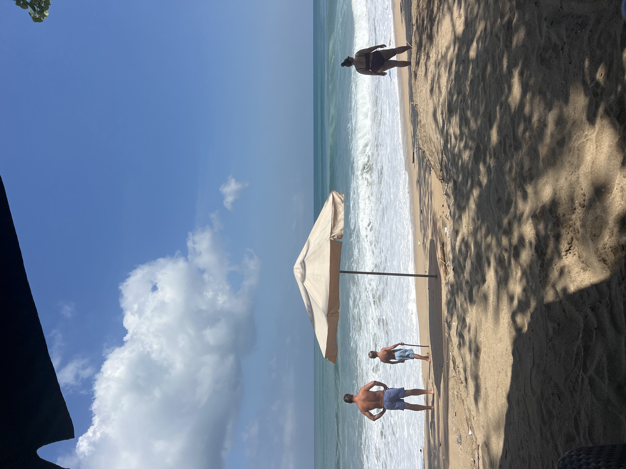The image shows a beach scene in Kuta, Indonesia. In the foreground, the sandy beach is partially covered by shadows from overhead foliage. In the middle ground, a light-colored beach umbrella stands on the sand, with waves crashing on the shore. Three individuals are present: a man and a boy stand near the water's edge, facing the ocean, with the man's hands on his hips and the boy holding a stick. Further down the beach, a woman in a dark bikini stands with her back to the camera, also facing the ocean. The sea is a vibrant blue-green, with white foam from the breaking waves. The sky above is a clear blue with scattered white clouds. The overall atmosphere is one of a sunny day at the beach.