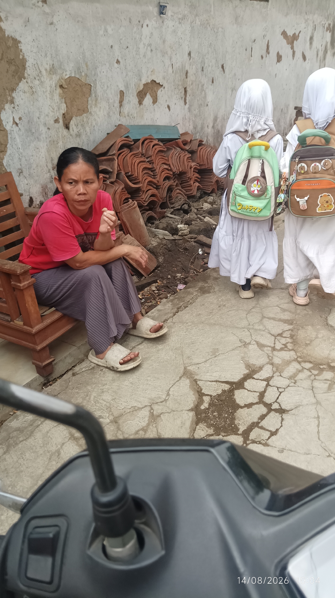 A woman in a pink t-shirt and grey pants sits on a wooden chair. She is looking to her right with a serious expression. She is wearing white sandals. Behind her, a stack of red roof tiles is piled against a weathered, white wall. Two children in white hijabs and dresses stand on the right side of the frame, facing away from the camera. The child on the left wears a green and beige backpack with a cartoon character, and the child on the right wears a brown backpack with cartoon characters and a zipper. The ground is made of cracked concrete. In the foreground, a motorcycle's handlebars and mirror are visible, suggesting the photo was taken from the perspective of someone on a motorcycle. A date stamp "14/08/2026 10:04" is present in the bottom right corner. The scene appears to be in an outdoor area, possibly a residential or construction site, in Tasikmalaya, Indonesia.