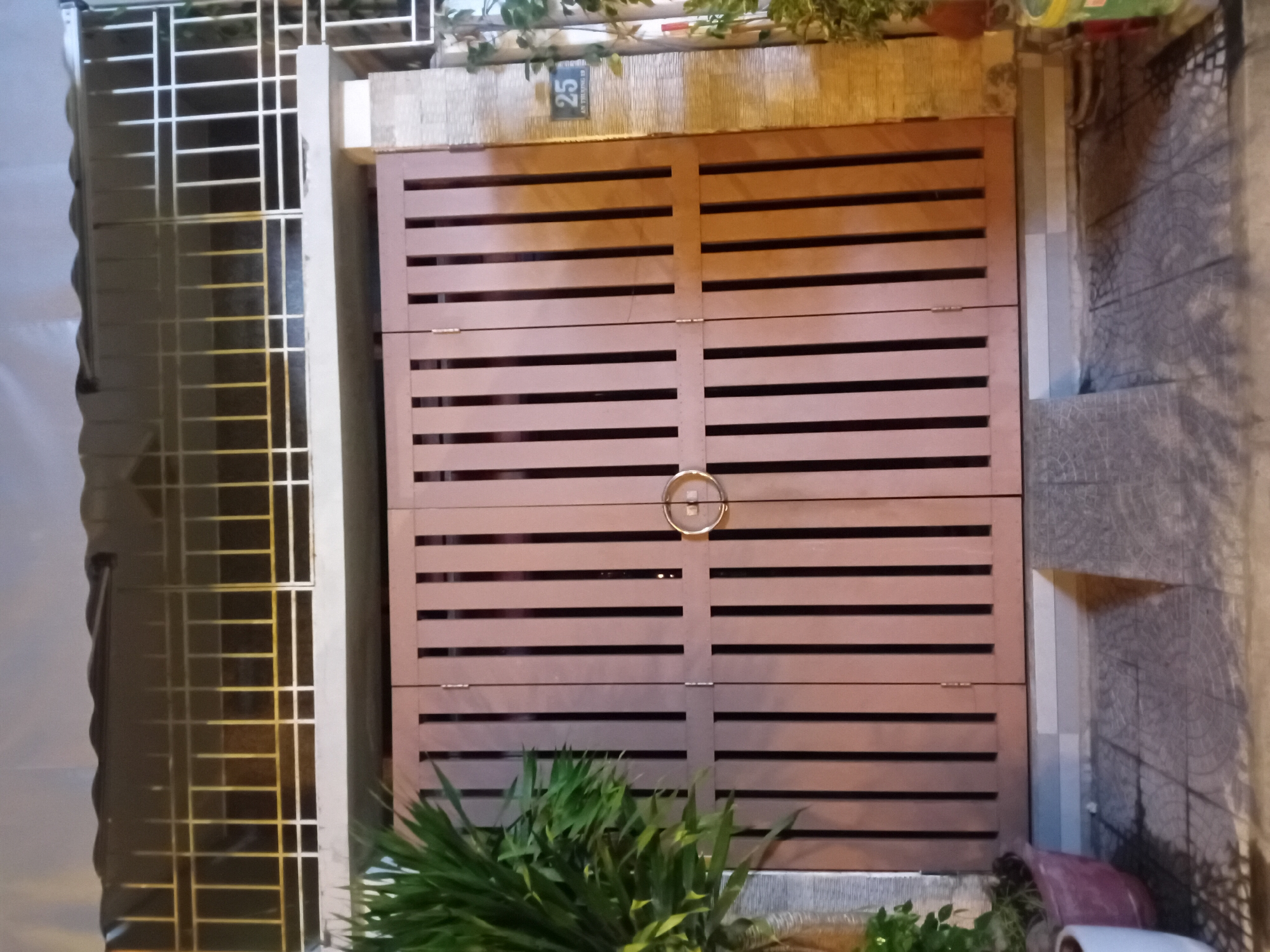 The image shows a closed, light brown wooden gate with horizontal slats, featuring a circular metal handle in the center. The gate is flanked by a white metal security grate on the left and a tiled wall on the right. Above the gate, a bamboo-patterned wall has a sign with the number "25" on it, along with some Vietnamese text. Lush green plants are visible in the foreground, partially obscuring the bottom of the gate and the ground. The scene appears to be outdoors, likely in a residential or commercial setting, given the presence of the gate and security grate. The lighting suggests it might be evening or nighttime, with artificial lights illuminating the area. The overall impression is one of a secured entrance to a property in Đà Nẵng, Vietnam.