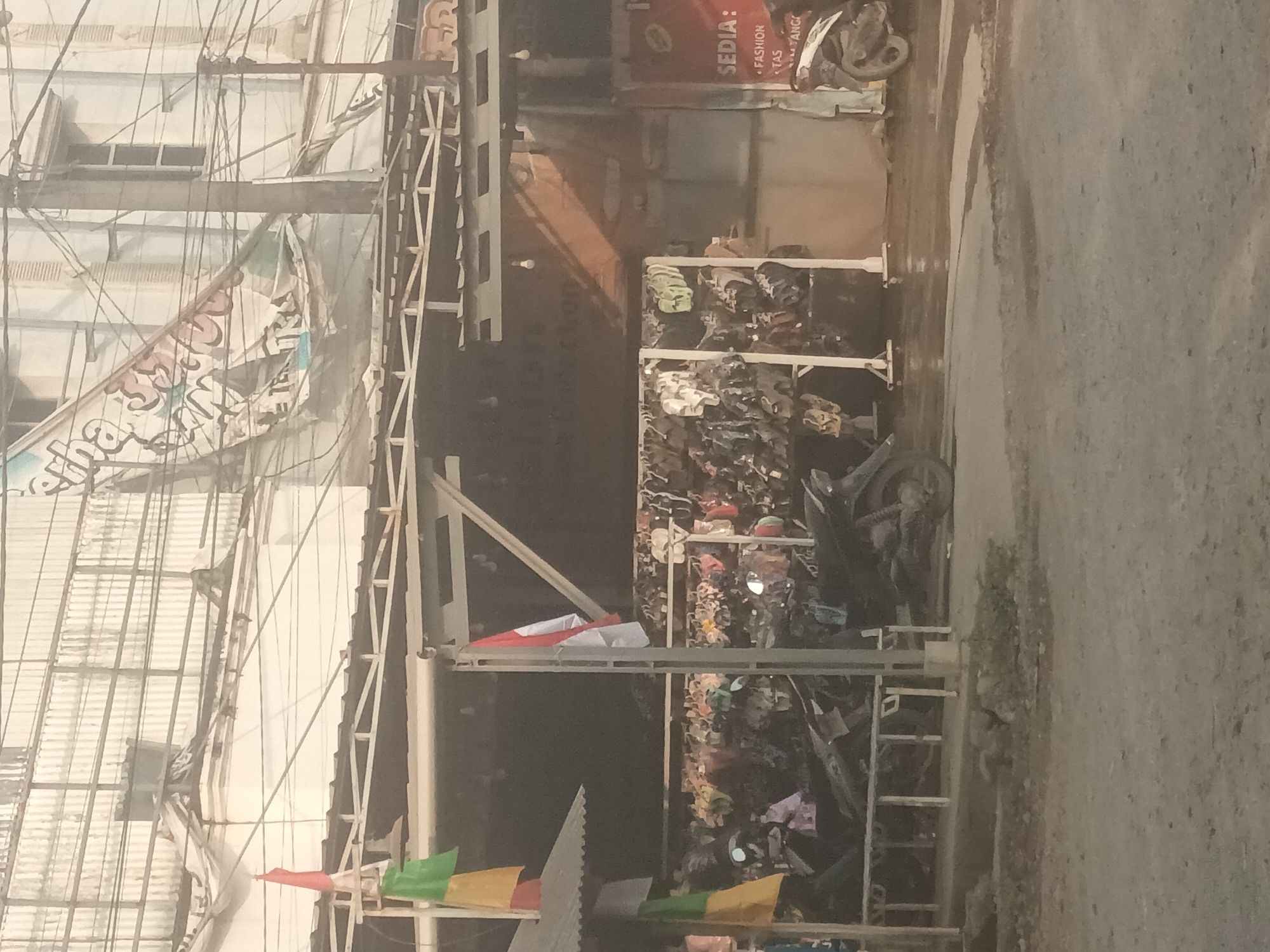 The image depicts a street scene in Mulyo Asri, Indonesia. In the foreground, a dirt and asphalt road is visible, with some overgrown grass patches. Behind the road, a row of motorcycles is parked under a covered structure, which appears to be a marketplace or a shop awning. The structure is supported by metal poles, and hanging from it are various items, possibly clothing or accessories displayed for sale. Further back, a building with a corrugated metal roof is visible, with electrical wires strung across it. A banner with Indonesian text and possibly graphics is draped across the upper part of the building, appearing somewhat tattered. An Indonesian flag is also prominently displayed on a pole near the covered market area. The overall atmosphere suggests a busy commercial area, likely in the late afternoon or early evening, given the lighting conditions.