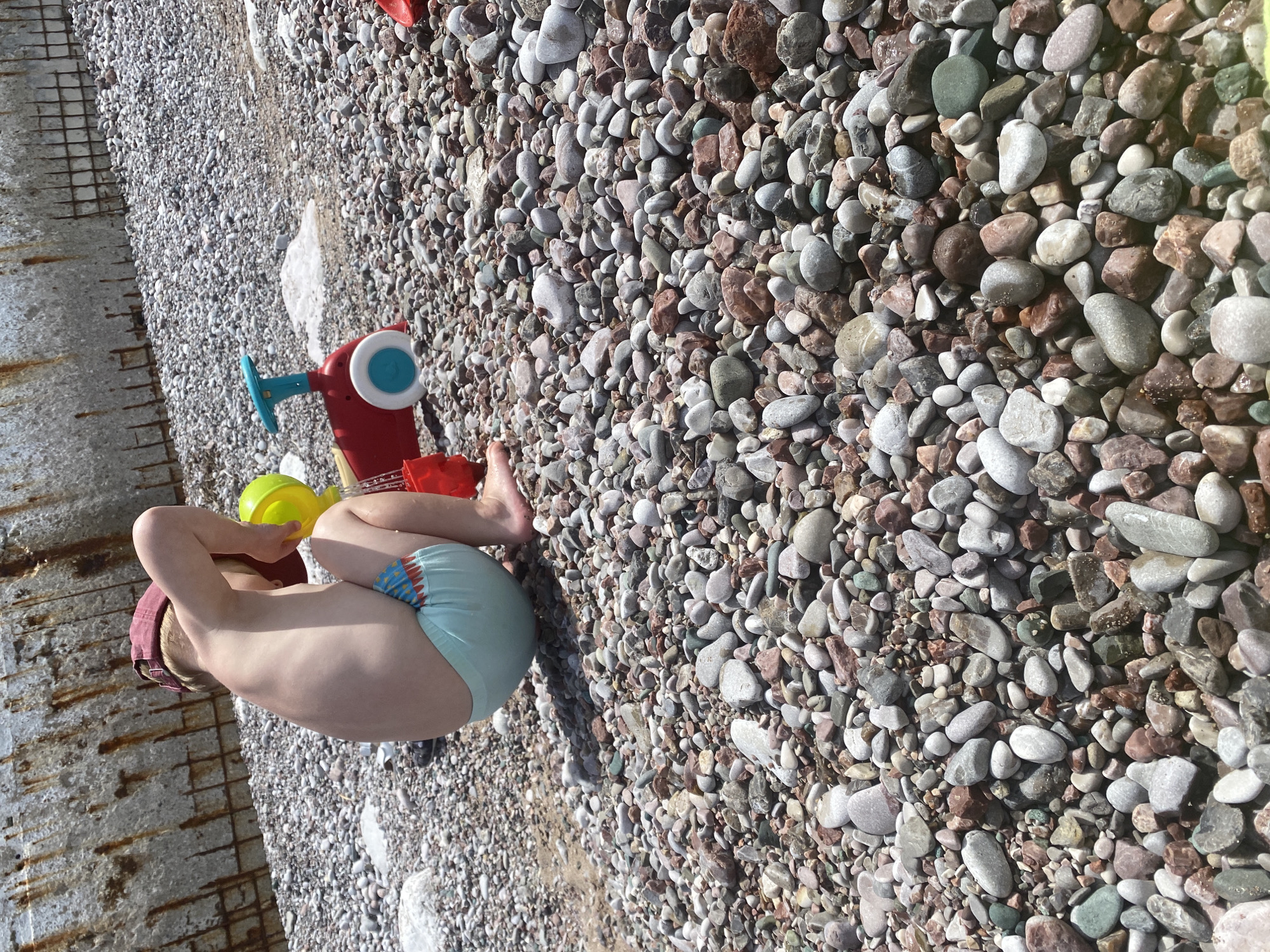 A young child is playing on a rocky beach next to a concrete wall. The child is wearing light blue swim trunks and a maroon cap. The child is playing with a yellow watering can and a red plastic toy. The beach is covered in pebbles and stones. The location is Rafailovići, Montenegro.