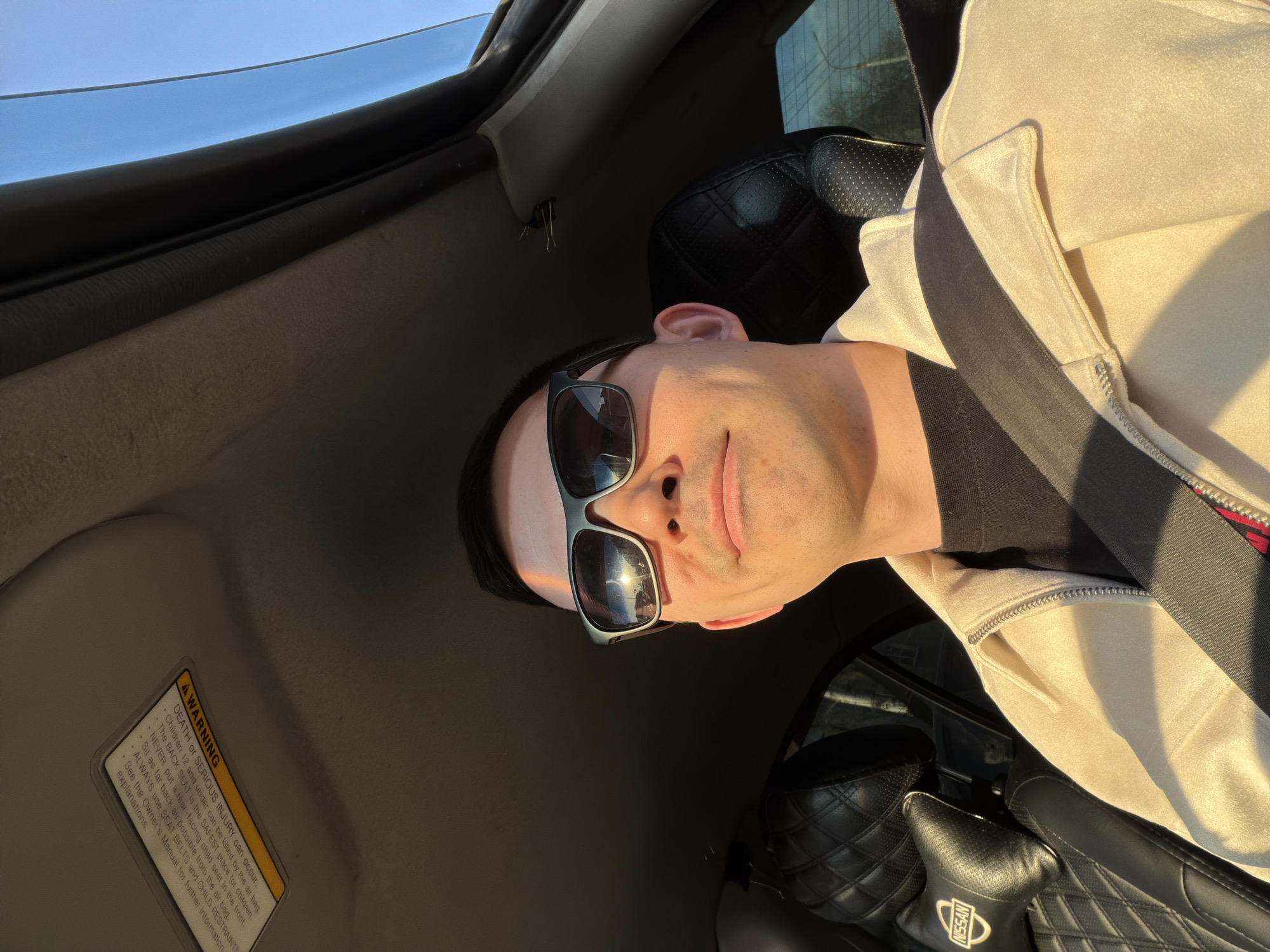 The image shows a man wearing sunglasses and a seatbelt in a car. The interior of the car is visible, including a sun visor with a warning label, quilted leather seats with a Nissan logo, and a partially open sunroof. The man is wearing a light-colored jacket over a dark shirt. The location is unknown and cannot be determined from the provided image.