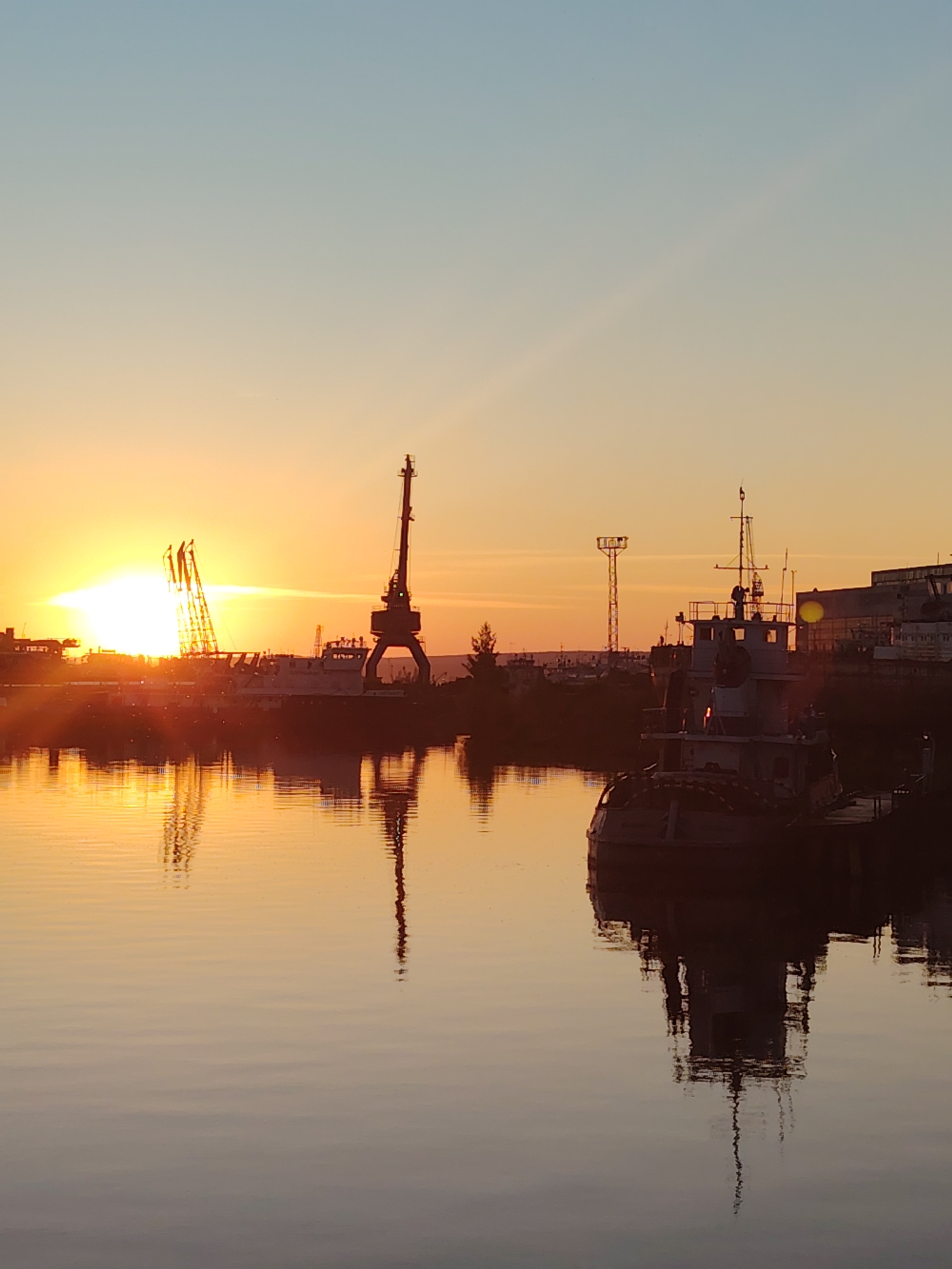 The image depicts a sunset over the Kama River in Chaikovsky, Russia. Silhouetted against the orange and blue sky are industrial cranes and boats docked in a harbor. The setting sun casts a warm glow on the water, creating reflections of the structures. In the foreground, the calm water of the river occupies the lower half of the frame, with ripples distorting the reflections of the objects on the shore. The sky transitions from a pale blue at the top to a vibrant orange near the horizon, where the sun is partially obscured by clouds and industrial elements. Several cranes, one large gantry crane and others that appear to be jib cranes, stand tall along the waterfront. Two boats, one a larger vessel with a superstructure and the other a smaller boat in the foreground, are visible. The overall scene conveys a sense of industrial activity at the end of the day, with the natural beauty of the sunset providing a dramatic backdrop.