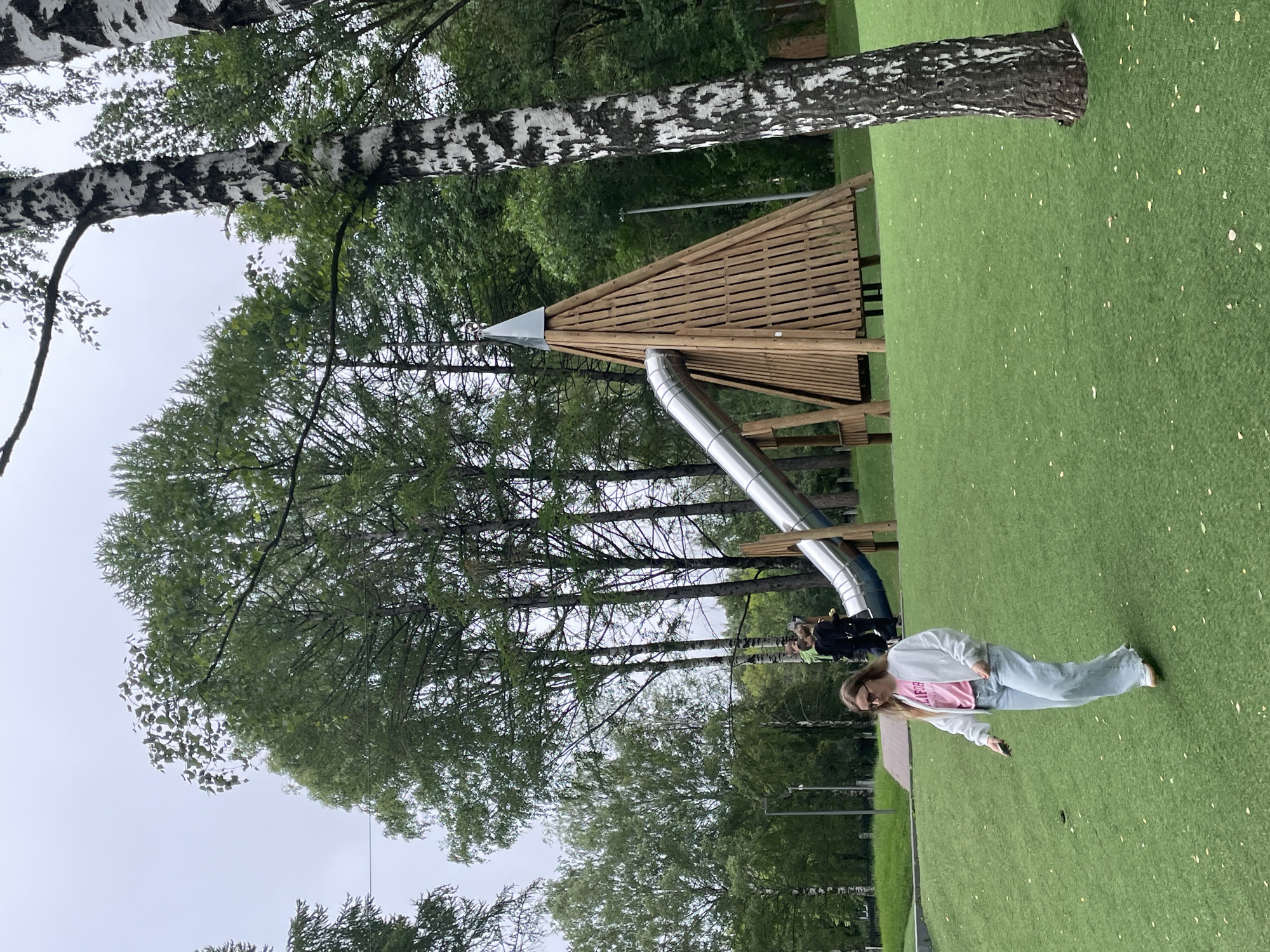 A wide-angle, eye-level shot captures a playground with artificial turf in Kirov, Russia. In the foreground, a young woman with blonde hair, wearing a white jacket over a pink t-shirt and light blue jeans, walks across the green turf, looking down at her phone. In the mid-ground, a large, silver slide descends from a conical wooden play structure topped with a silver pyramid. Behind the structure, several tall birch trees with distinctive black and white bark are visible, along with a stand of other deciduous trees. The sky is overcast and grey. Two people are partially visible at the top of the slide, appearing to be looking down. The overall scene suggests a public park or recreational area.