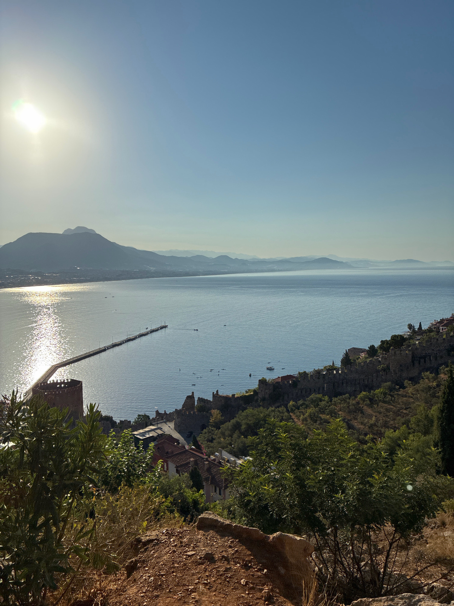 The image captures a scenic view of Alanya, Turkey, from a high vantage point overlooking the Mediterranean Sea. In the foreground, lush green foliage and rocky terrain dominate, hinting at a natural, elevated position. The midground reveals the ancient Alanya Castle walls, with their distinctive red brick towers, curving along the coastline. Below the castle, a collection of terracotta-roofed buildings is nestled into the hillside, indicative of the city's architecture. A long pier extends out into the calm, blue sea, dotted with several small boats. In the distance, hazy mountains form a picturesque backdrop, their silhouettes softened by the morning or evening light. The sun is bright and positioned in the upper left corner of the frame, casting a shimmering reflection on the water and creating a sense of warmth and tranquility. The sky is a clear, pale blue, with no clouds visible, further enhancing the peaceful atmosphere of the coastal city.