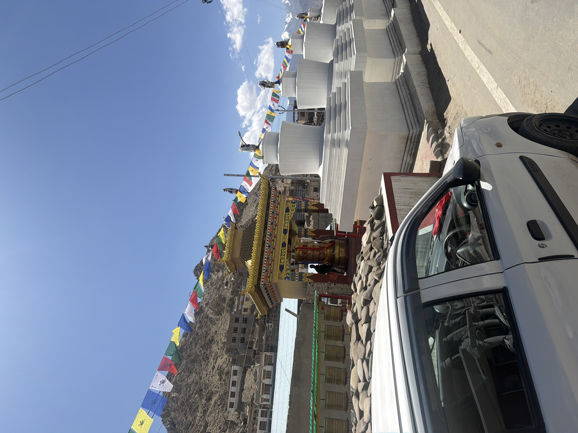 In the city of Leh, India, a white car is parked on the right side of a paved road. The car is facing towards the left of the frame, with its front fender and side visible. In the background, a Buddhist monastery complex is situated on a hillside under a clear blue sky. Prayer flags in various colors are strung across the scene, fluttering in the breeze. Several white stupas, characteristic Buddhist structures, are lined up on the right side of the monastery. The monastery itself features a golden roof and ornate decorations. Prayer wheels are visible in front of the monastery, some of them lined with stones. Buildings are clustered on the hillside behind the monastery, with a rocky, mountainous terrain forming the backdrop. The sun is bright, casting shadows on the road and the structures.