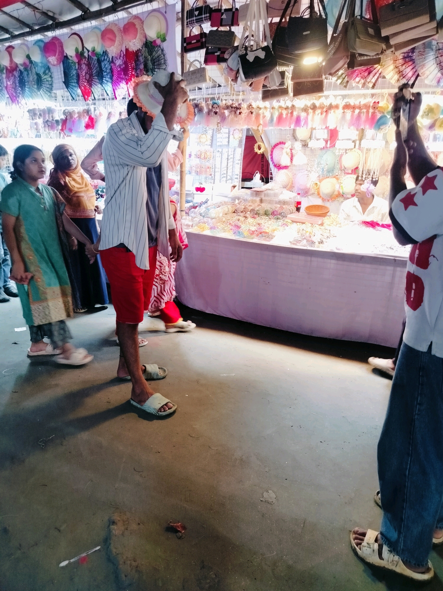 This is a medium-full shot of a bustling market stall in Dhaka, Bangladesh, likely a street market or a temporary setup during a festival. The stall is laden with various accessories, including colorful hats, fans, jewelry, and handbags. The scene depicts people browsing and interacting with the merchandise.
In the foreground, a person wearing a white and black striped shirt and red shorts is holding a hat, examining it. They are wearing light-colored sandals. To the left of this person, a young girl in a green outfit and a woman in a yellow hijab are also present, seemingly looking at the merchandise. Further back, a man in a white shirt is visible behind the counter, attending to the stall. Another person, whose back is to the camera and wearing a white t-shirt with red star patterns and blue jeans, is also visible on the right side of the frame, seemingly holding something up, possibly for examination. The ground appears to be a concrete surface, with some debris scattered around. The stall is covered by a white tarp, and the background is filled with hanging accessories, creating a vibrant and crowded atmosphere. The lighting suggests it could be daytime, possibly late afternoon given the shadows.