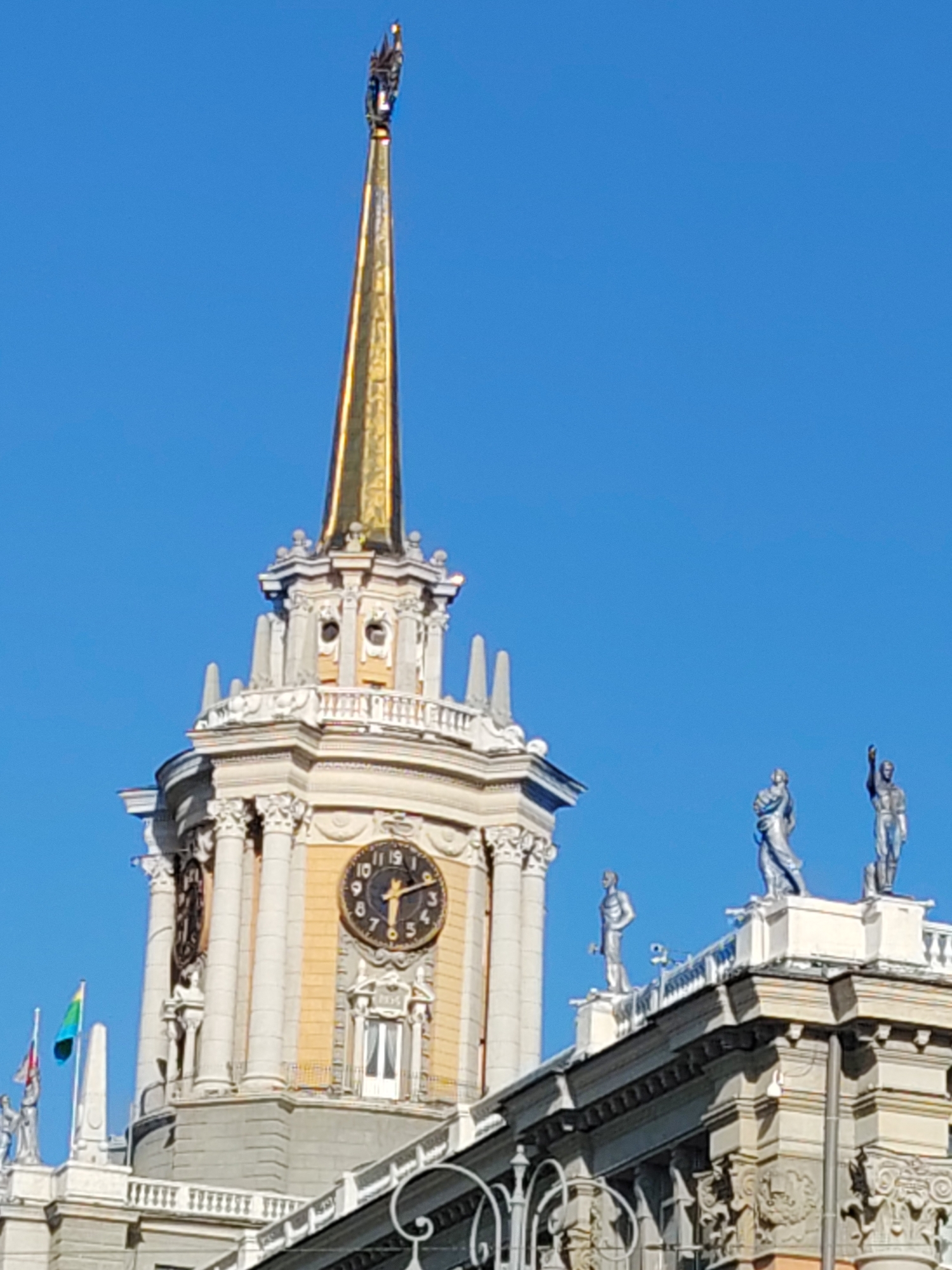 The image captures a close-up view of the Yekaterinburg City Administration building's clock tower and adjacent rooftops against a clear blue sky. The dominant feature is the ornate clock tower, topped with a gilded spire and a detailed sculpture at its apex. Two clocks are visible on the tower's facade, with one clearly showing the time as approximately 10:10. The architecture is classical, with columns, decorative moldings, and multiple levels. Several statues of human figures in classical attire are positioned on the rooftops of the main building, one with an arm raised. Two flags, one with green and yellow bands and another with red, white, and blue, are visible flying from a flagpole on the left side of the tower. The scene is bathed in sunlight, casting shadows that highlight the architectural details.