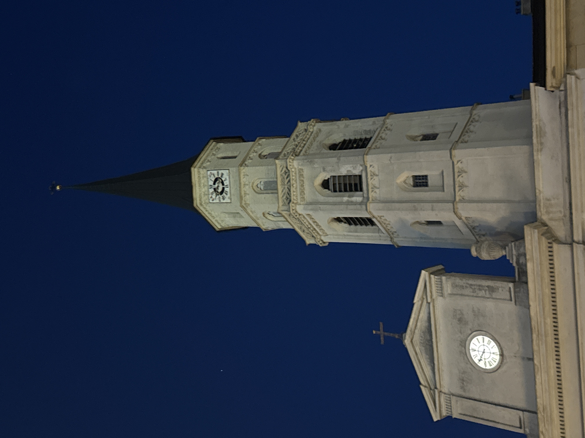 This is a photograph of the Stephansdom, also known as St. Stephen's Cathedral, in Vienna, Austria, taken at night against a deep blue sky. The image focuses on the cathedral's tower and a section of its facade. The tower is tall and slender, with a dark, pointed roof. It features multiple levels with ornate architectural details, including arched windows and decorative carvings. A clock face is visible on one of the upper levels of the tower. To the left of the tower, a portion of the cathedral's roofline and a smaller structure topped with a cross are visible. This structure also has a clock face, displaying Roman numerals. The lighting suggests artificial illumination of the building, creating a stark contrast with the dark sky. There are no people or active interactions depicted. The overall scene is one of architectural grandeur and stillness in the nighttime environment.