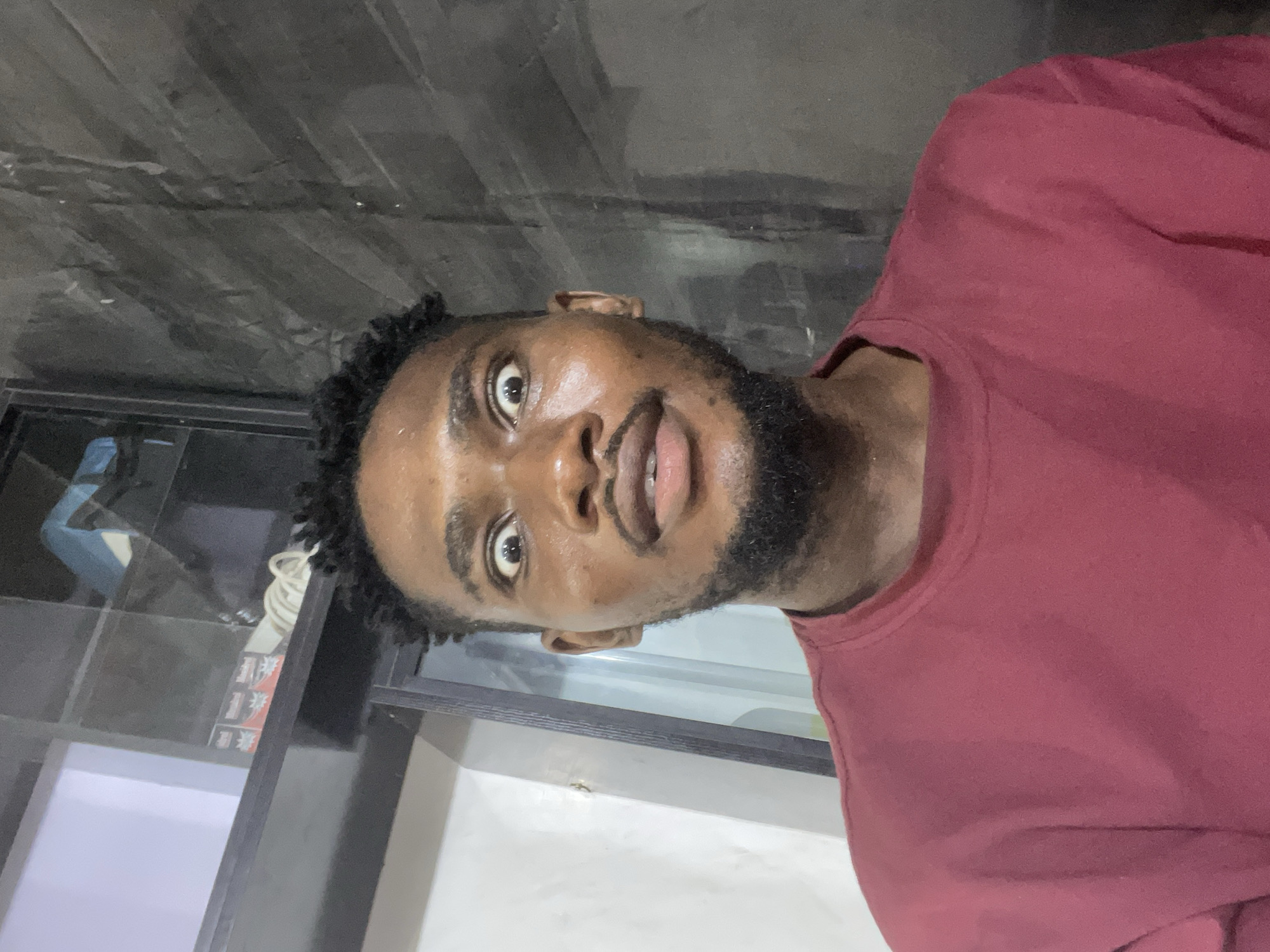 A close-up, slightly tilted, indoor portrait of a dark-skinned man with a trimmed beard and dark hair. He is wearing a maroon t-shirt. He is looking directly at the camera. The background includes a dark gray wall and a white surface with some items on it. The location is likely indoors, but there is no clear indication that it's in Unguwar Gabas, Nigeria.