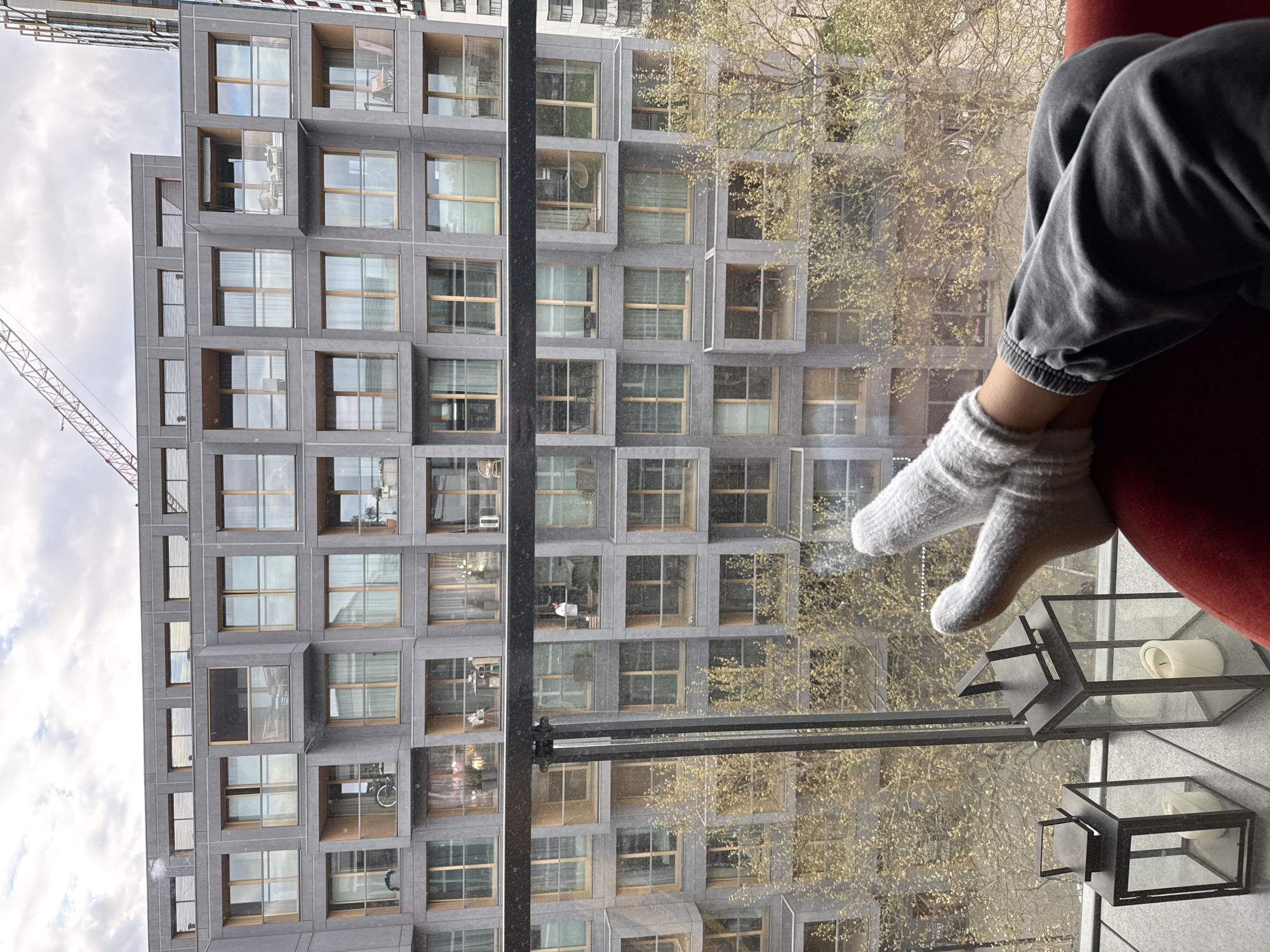 The image, captured in Warsaw, Poland, shows a relaxed indoor perspective looking out onto a modern urban environment. In the immediate foreground, a person's lower legs and feet are visible, resting comfortably. The person wears grey, slightly wrinkled sweatpants and light grey or white knitted socks, with their legs positioned against or on a piece of red furniture, likely an armchair or sofa. To the lower right, two decorative, dark-framed lanterns with glass panels, each containing a white candle, are visible, placed on what appears to be an interior ledge or balcony.
Beyond the window, a large, multi-story modern building dominates the view. Its facade features a geometric grid of dark, possibly concrete or stone, panels and numerous rectangular windows. Many of these windows are recessed, forming small balcony or loggia spaces. The architecture suggests a contemporary residential or commercial structure. Between the viewer's window and the building, the branches of several trees are visible, adorned with sparse, light green or yellowish leaves, indicating springtime.
The sky on the left side of the image is partly cloudy, with white and grey clouds. A construction crane is partially visible on the far left, extending upwards behind the modern building, suggesting ongoing development in the area. The scene conveys a sense of quiet observation from an elevated position. There is no discernible text visible in the image.