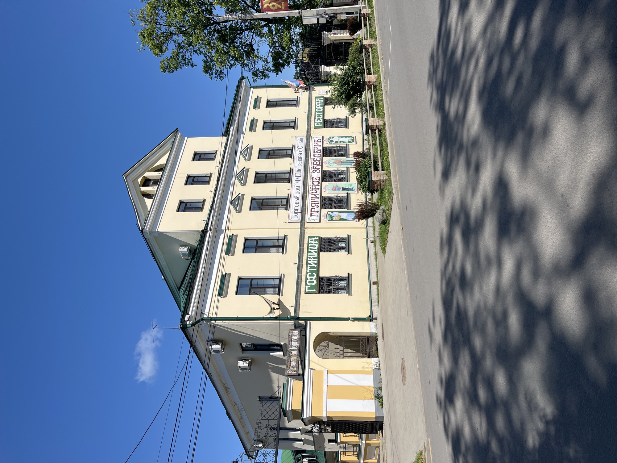 A light yellow, three-story building with a green roof is situated in Kladovitsy, Russia. The building features white trim around the windows and along the roofline. The front facade is adorned with several signs. One sign, above an arched entryway, reads "Гостиница" (Hotel). Another sign, in a smaller font, states "Торговый домъ ММПлешанова с-ми" (Trading house of M.M. Pleshano and company). Below this, a larger sign declares "ПРЯНИЧНОЕ ЗАВЕДЕНИЕ" (Gingerbread establishment), with decorative paintings of people in traditional attire on the wall beneath it. To the right of this, a sign in green letters reads "РЕСТОРАНЪ" (Restaurant). The building has numerous windows, many with dark-colored frames. There is a decorative wrought-iron fence in front of the building. To the left of the main entrance, a smaller section of the building with a yellow and white striped facade is visible, also with a gate. A tall green tree stands to the right of the building. The building is located on a street with a paved road and sidewalk. The sky is clear and blue, with a few wispy clouds. Shadows of tree branches are cast across the road and sidewalk, indicating a sunny day.