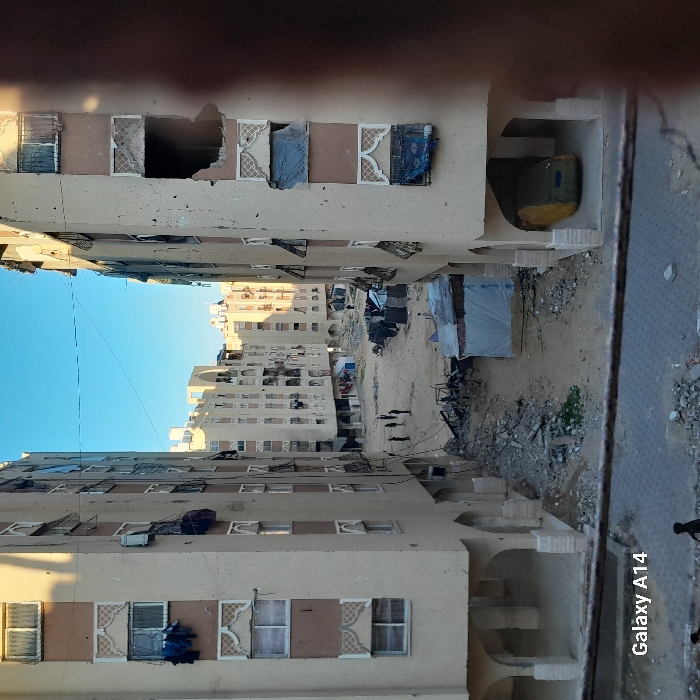 This image depicts a damaged residential area in Khan Yunis, Palestinian Territory. Two multi-story, light-colored concrete apartment buildings flank the foreground, framing a central unpaved area. Both buildings exhibit extensive damage, including numerous impact holes on their facades, shattered windows, and missing sections of walls. The building on the right has a large, multi-story irregular void where a section of its facade has collapsed. Some windows are covered with tarpaulins or fabric, while others have laundry hanging from them. The ground floors feature open arched passages; one on the right building contains a green tarp-covered vehicle.
The unpaved ground between the buildings is covered in heavy rubble, concrete fragments, and debris. Scattered among the debris are several makeshift structures, appearing to be temporary shelters constructed from tarpaulins and corrugated metal, with a prominent white tarp structure in the mid-ground. Approximately five to seven individuals, including adults and possibly children, are visible standing or walking on the dirt path in the mid-ground. Another partial figure is visible at the very bottom edge of the frame. The sky is clear and blue, with sunlight casting angular shadows across the scene, indicating daytime.