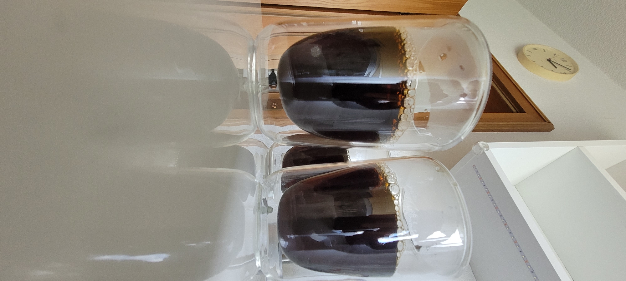 The image shows a close-up, low-angle shot of three clear glass mugs filled with dark liquid, likely coffee, resting on a reflective white surface. The mugs have a double-walled design, and the liquid inside has a foamy top layer. In the background, on a white wall, a round, light-yellow clock with black markings and hands is visible. The clock shows the time to be approximately 11:50. To the left of the clock is a wooden door frame. Above the mugs, a white shelf with LED lights is partially visible. The overall scene appears to be an indoor setting, possibly a kitchen or dining area. The image does not provide any specific information about the location in the city of Heusenstamm, Germany. There are no people visible in the image.