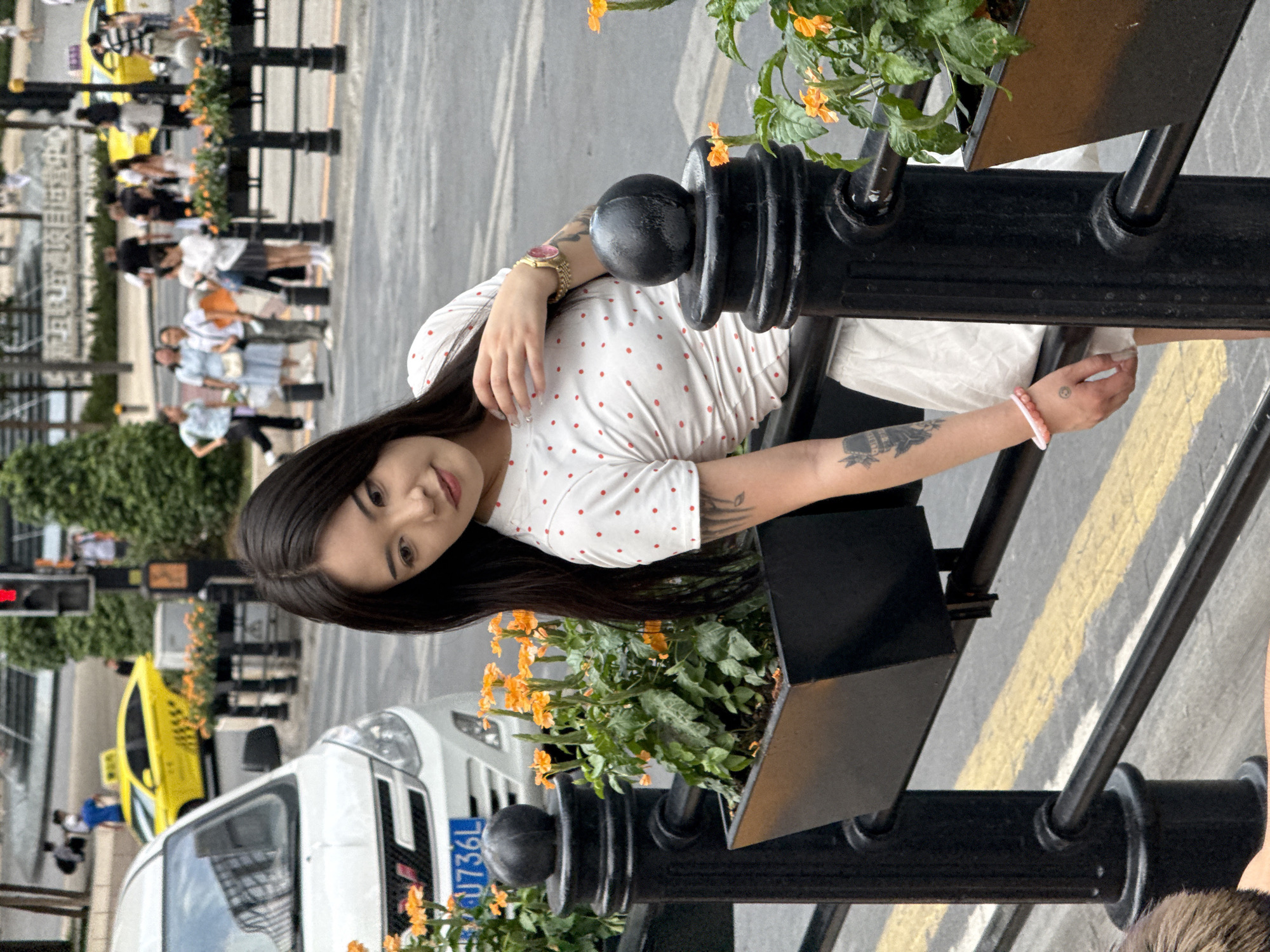 A young East Asian woman with long dark hair leans on a black metal railing in Yuzhong District, China. She is wearing a white t-shirt with red polka dots and a white skirt. She has tattoos on her left arm and a pink bracelet on her left wrist. The railing is in front of a black planter box with orange flowers. In the background, a white van is parked, and a yellow taxi is driving on the street. Several people are walking on the sidewalk in the distance. The environment appears to be an urban street scene.