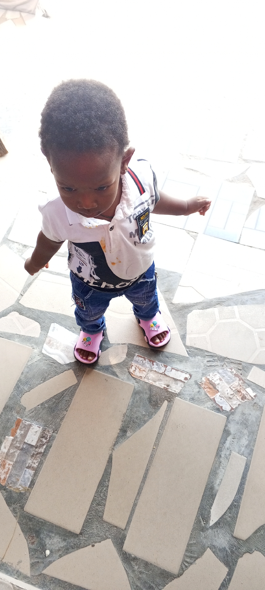 An overhead shot shows a young Black child wearing a white polo shirt with black and red accents, and blue jeans. The shirt has several patches and graphics, including one that says "NO. P.5" and another with "METAL". The child is also wearing pink sandals. The child is standing on a tiled floor with a mosaic pattern of beige and grey tiles. The tiles are irregularly shaped and fitted together with grey grout. Some of the tiles appear to be broken or chipped, revealing concrete underneath. The lighting suggests it is daytime, possibly sunny, casting shadows on the ground. The background is blurred but appears to be an outdoor setting. There is no clear indication of the specific event, but the child appears to be standing and looking down, possibly at the ground. The location is identified as Jalingo, Nigeria.