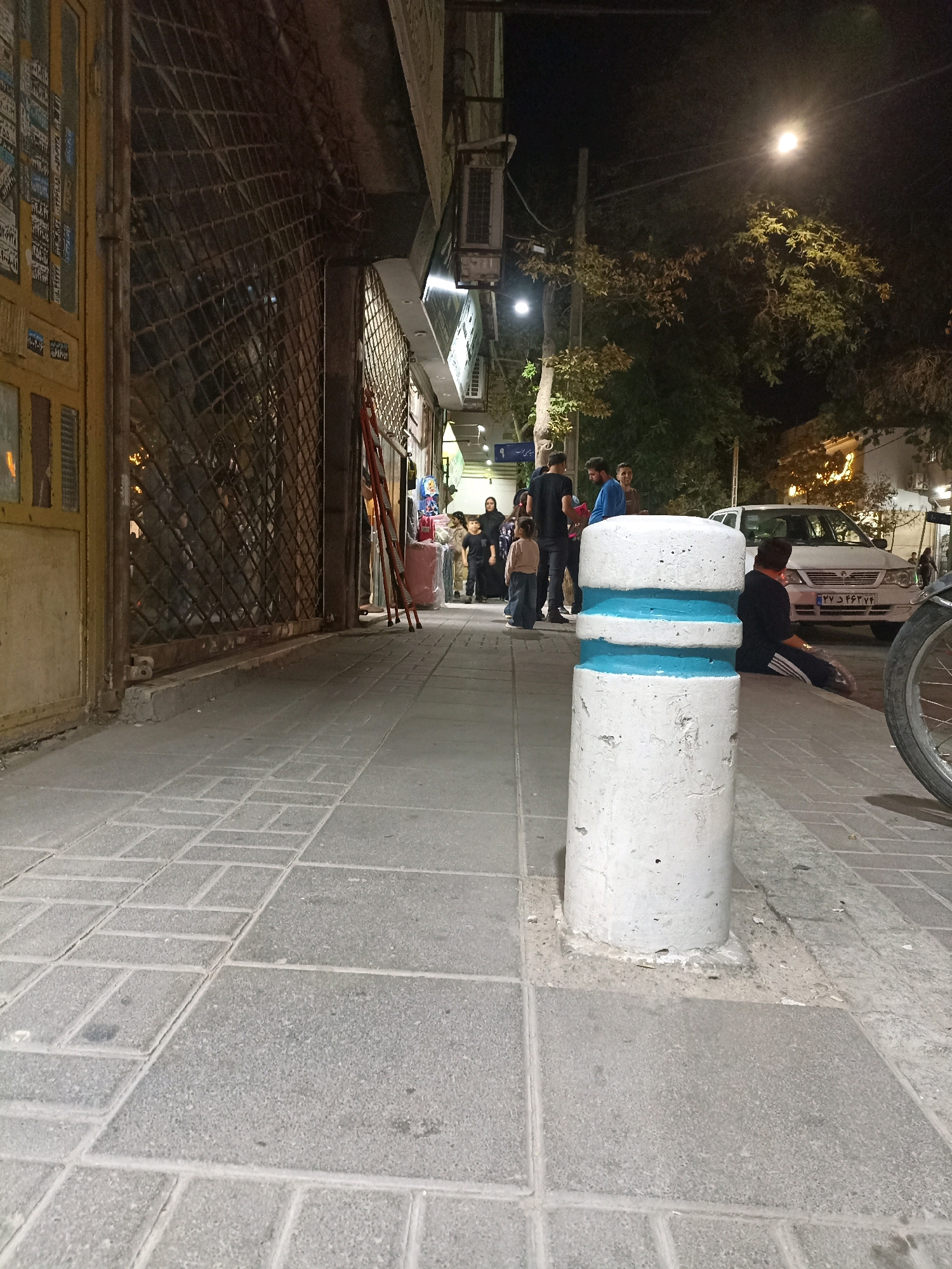 The image captures a street scene at night in Mashhad, Iran. In the foreground, a textured concrete bollard painted with two blue bands stands on a sidewalk paved with rectangular gray stones. To the left, the facade of a building with a yellow door and a metal security grate covers a storefront. Further down the street, several people are visible. A group of individuals, including adults and children, are gathered near the storefronts. A white sedan is parked on the street to the right, and a motorcycle wheel is also visible in the far right of the image. The lighting suggests an urban environment with artificial lights illuminating the area.