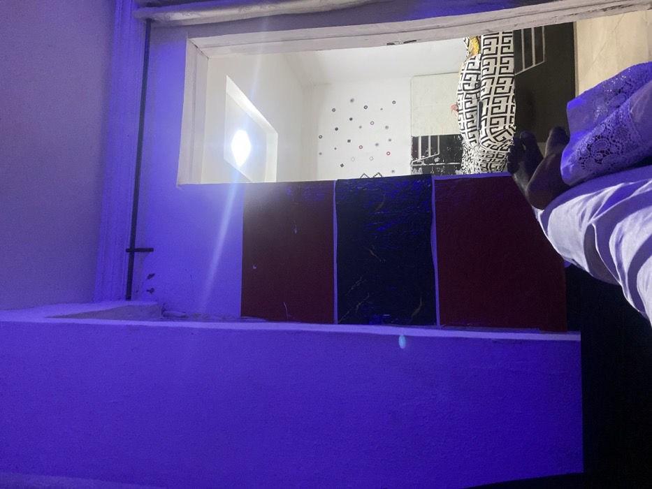 The scene depicts an interior view within a residential building in Ibadan, Nigeria. The immediate foreground is illuminated by a pervasive blue artificial light. In the lower right of the frame, the legs of a person, clad in light-colored trousers, are partially visible, with a small, dark-furred animal resting upon them. A segment of the wall to the left of a doorway features three distinct rectangular panels: the top and bottom panels are reddish-brown, while the central panel is black with a textured, reflective surface. A dark, horizontal rod or curtain rail is mounted above the doorway, near the ceiling.
Through the doorway, a second, brightly illuminated room is visible. This room features white walls, a white recessed ceiling with a central square LED light fixture, and a bed covered with a black and white geometric patterned duvet. The wall above the bed is decorated with numerous small, multi-colored circular and square decals. No other individuals are discernible within the visible areas of either room.