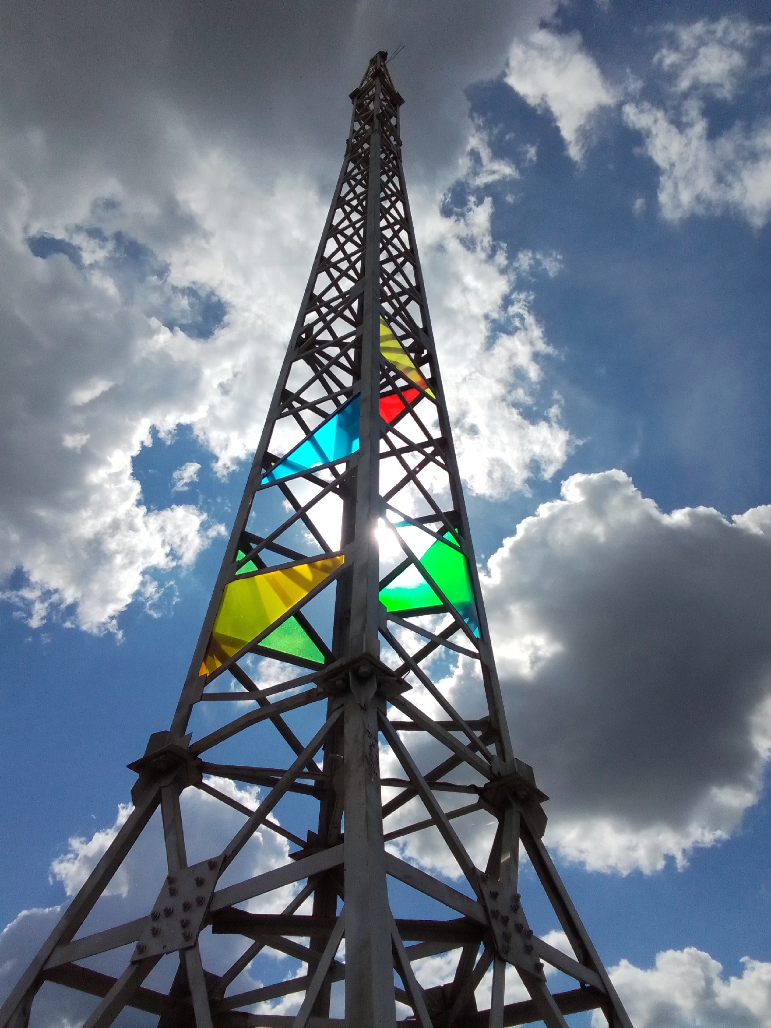An upward-angle, medium shot captures a tall, metallic lattice tower against a bright, partly cloudy sky. The tower is constructed of interconnected metal beams and is silhouetted against the sky. Several panels of colored translucent material—red, yellow, blue, and green—are fitted within the latticework at different levels. The sun shines brightly, with a lens flare visible where it passes through one of the blue panels. The sky is a vibrant blue, with a mix of fluffy white clouds and darker, more ominous gray clouds. The image appears to be taken from the ground, looking up at the tower.