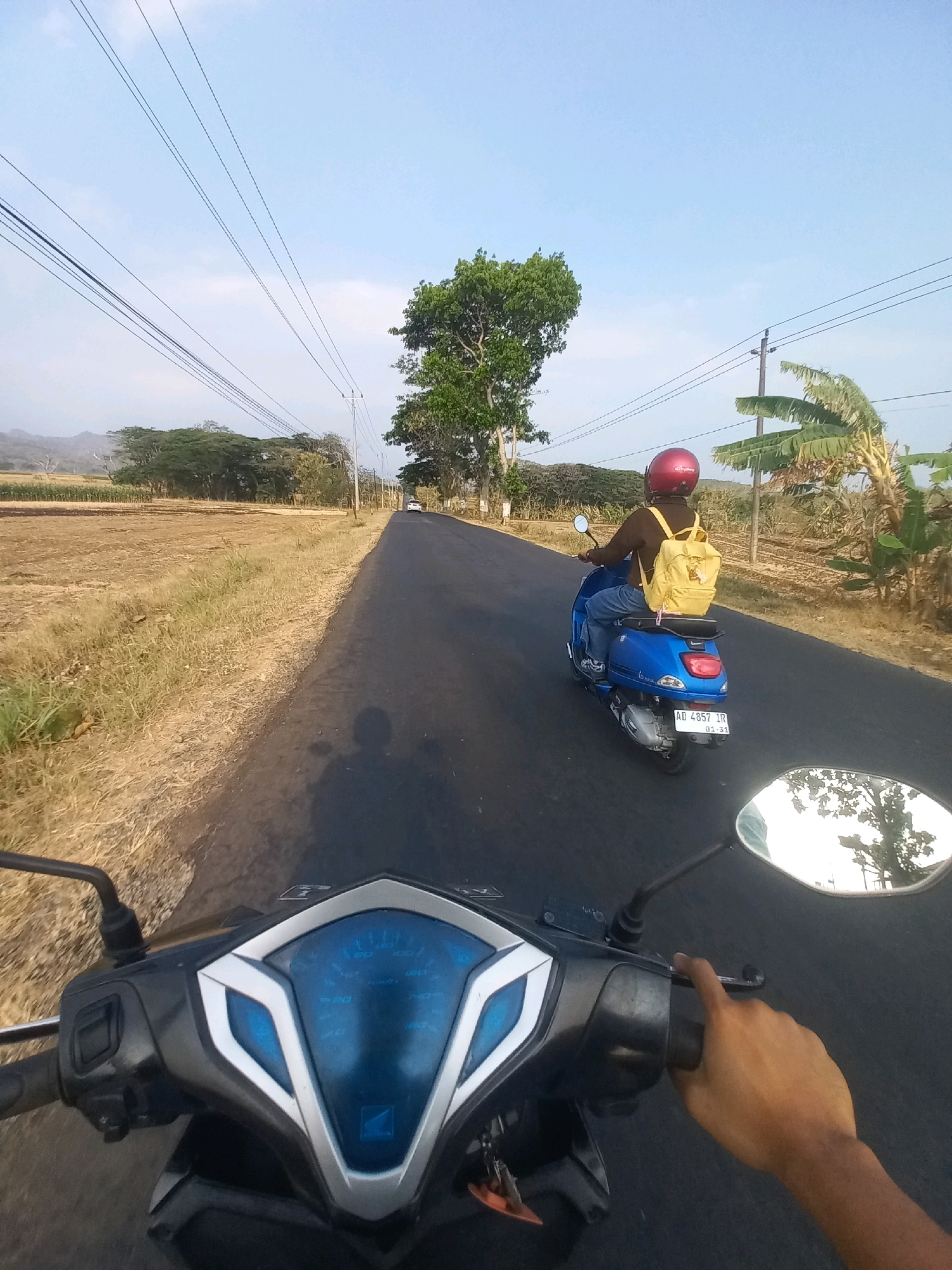 This is a first-person perspective video still of two people riding motorcycles down a paved road in Kedungombo, Indonesia. The foreground shows the handlebars, dashboard, and a hand of the rider of the first motorcycle. In the middle ground, a second person wearing a red helmet and a yellow backpack rides a blue Vespa scooter. The background features a rural landscape with dry fields, scattered trees, and hills under a clear sky. A power line runs overhead along the road.