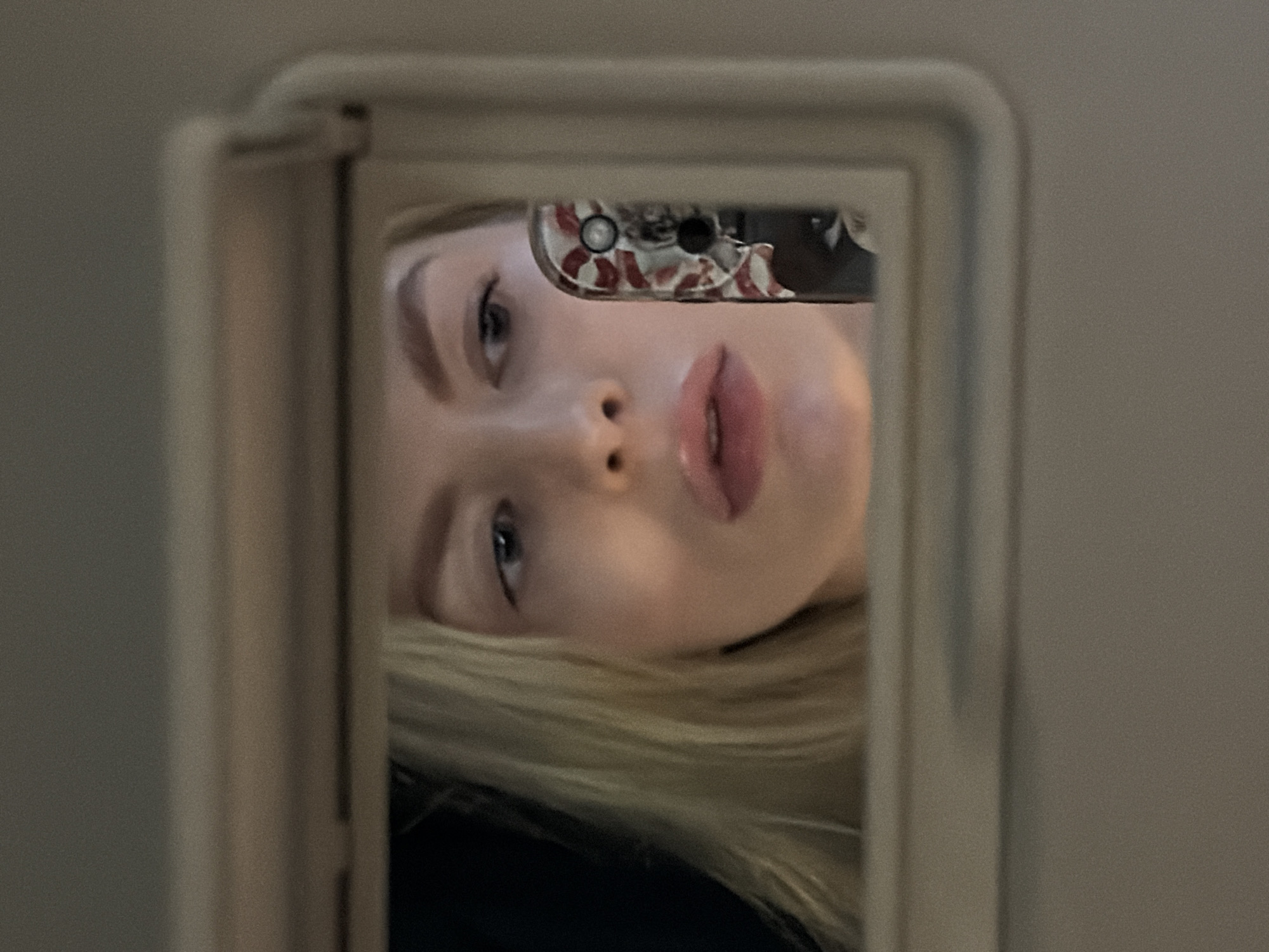 A close-up, frontal reflection shows a young Caucasian woman with blonde hair and light skin looking into a car's vanity mirror. Her mouth is slightly parted, revealing her pink lips. She is holding a smartphone, which is also reflected in the mirror. The phone case has a red, white, and black pattern. The background is dark, suggesting it is nighttime or the interior of a car with dimmed lighting. The mirror is framed by a beige visor. There is no discernible information about the specific location in Moscow, Russia, within the image itself.