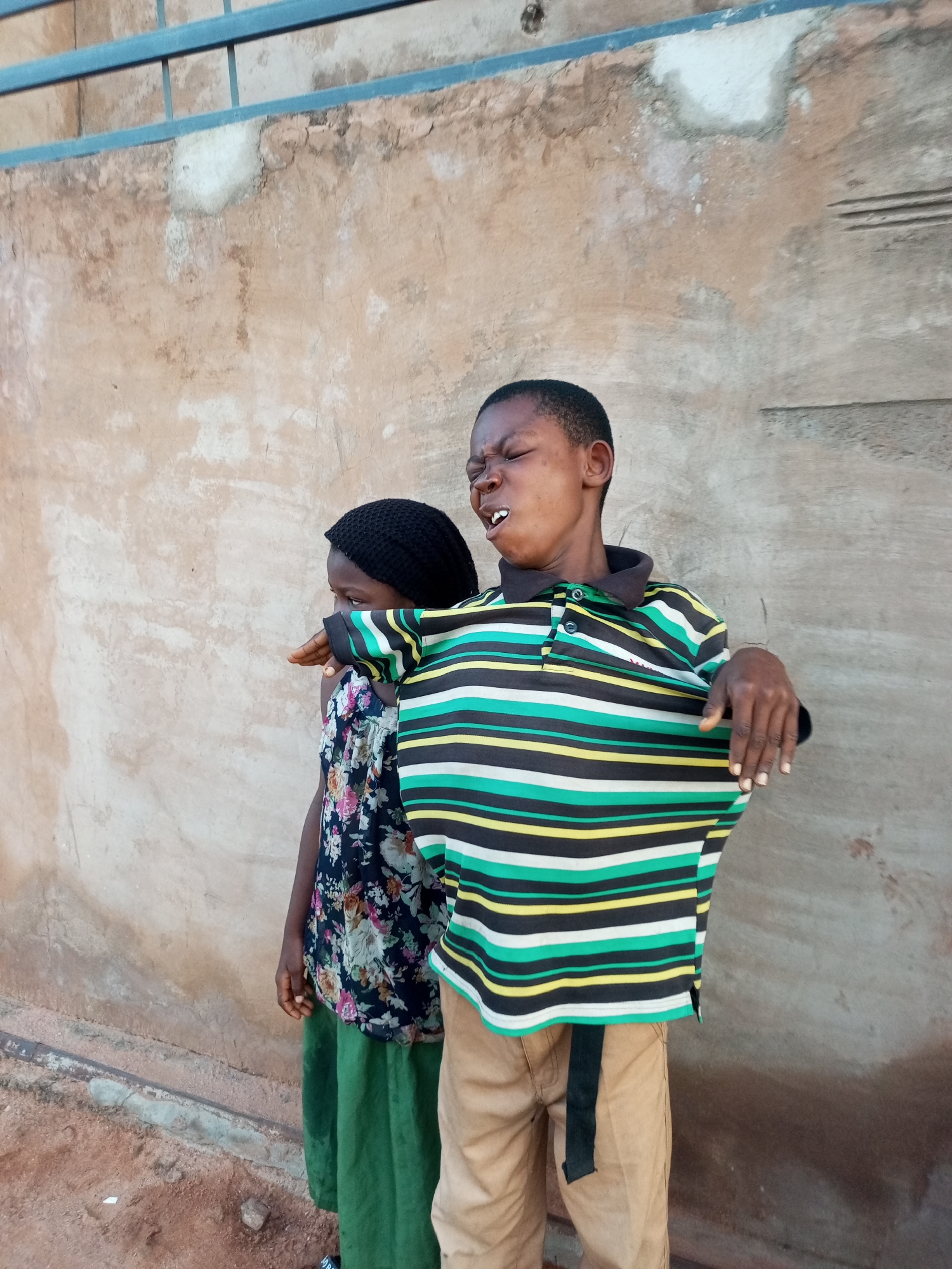 Two children stand against a textured, light brown wall. The child on the right, a boy, is in the foreground, facing slightly away from the viewer and looking upwards with his eyes closed and mouth open, showing his teeth. He wears a striped polo shirt with horizontal bands of black, white, green, and yellow, and tan pants with a black belt. His left arm is extended, with his hand resting on the shoulder of the child on the left. The child on the left, a girl, is partially obscured, facing away from the camera, looking towards her right. She wears a black head covering and a dark floral print dress with a green skirt. The wall behind them has a rough texture and appears to be made of stucco or concrete. Above the wall, a blue metal railing with horizontal bars is visible. The ground in front of the wall is reddish-brown dirt.