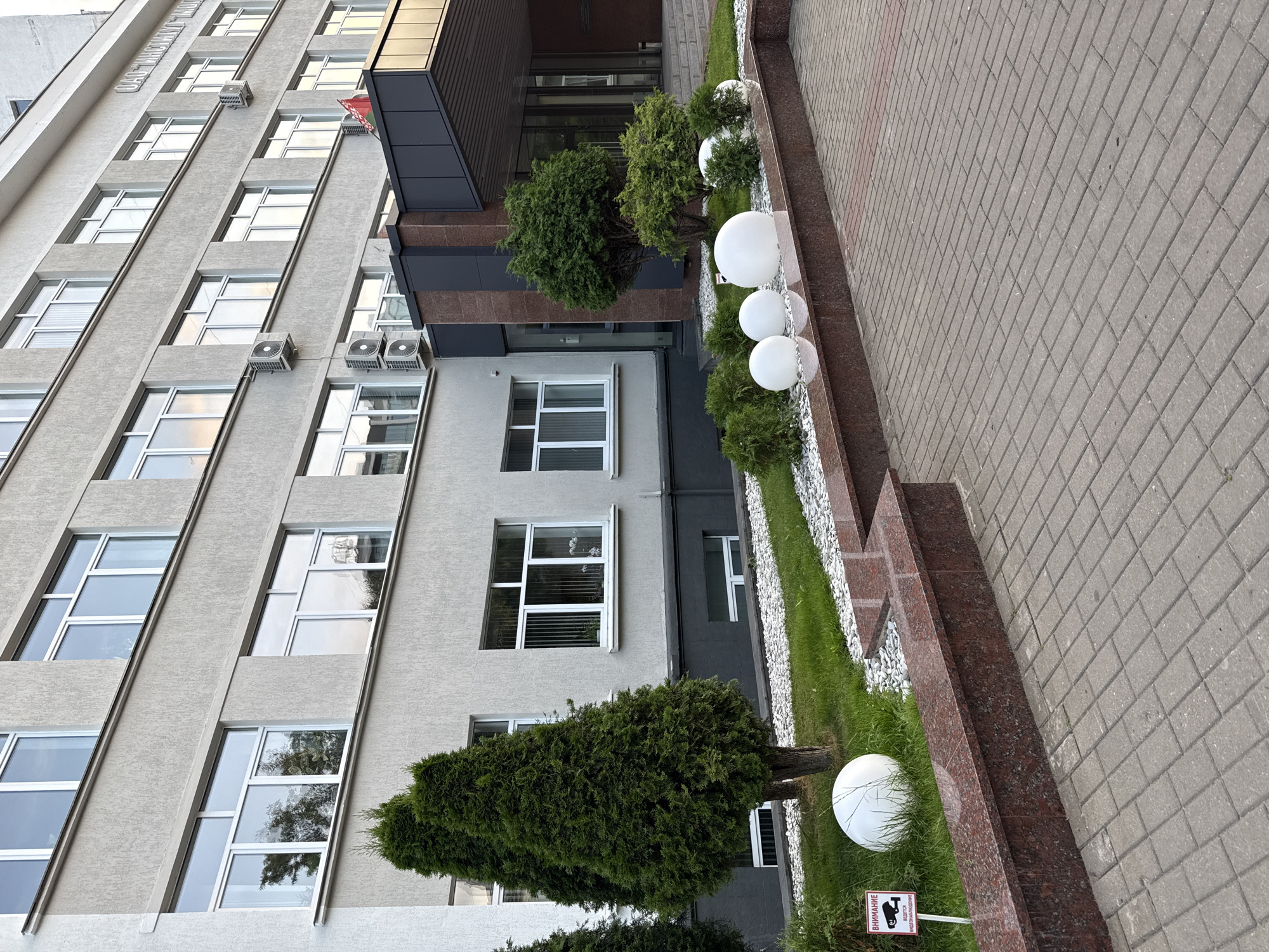 The image depicts the exterior of a modern, multi-story building, likely an office or administrative building, located in Mahilyow, Belarus. The facade is predominantly light gray with a textured finish, featuring numerous rectangular windows with white frames. Several air conditioning units are visible mounted on the exterior walls.
In the foreground, a paved area made of gray rectangular bricks leads towards the entrance of the building. Adjacent to the paved area is a raised flower bed with a reddish-brown granite border. This flower bed contains neatly trimmed green bushes and several large, white spherical decorative objects. Further into the flower bed, white gravel and grass are visible, interspersed with more green bushes and two tall, conical evergreen trees.
To the left of the evergreen trees, a small red and white sign with black text and an icon of a security camera is placed on a stake in the grass. The text on the sign appears to be in Cyrillic script and reads "ВНИМАНИЕ" (Attention) and "Ведётся видеонаблюдение" (Video surveillance is being conducted).
The building's entrance is recessed and features a dark gray canopy supported by dark brown columns. The entrance itself has large glass doors and windows. A flag, appearing to be red with a white emblem, is visible on a flagpole near the entrance. The overall environment suggests a well-maintained urban setting.