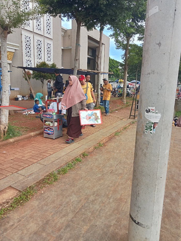 This image depicts an outdoor scene, likely a public or semi-public area in Singaparna, Indonesia, characterized by a mix of paved pathways, green spaces, and buildings. The time of day appears to be daylight, possibly late morning or afternoon, under a partly cloudy sky.
In the mid-ground, a woman wearing a pink hijab, a brown top, and a long brown skirt is walking from left to right. She holds a white board with a colorful, hand-drawn illustration. Adjacent to her, a man pushes a small, silver, two-wheeled cart labeled "SINGKA FRUIT," which contains various food items or fruit preparations. Several other individuals are scattered in the background; some men are standing and conversing, one notably wearing a yellow T-shirt and a red baseball cap, and another in a yellow T-shirt and a grey cap. Further back, under a dark fabric canopy, a small group of people, including a child, are seated on the ground, suggesting a small market stall or gathering spot.
The background is dominated by a light-colored building with distinctive architectural features, including windows or wall panels adorned with intricate, geometric patterns. This structure suggests a mosque or a public building. Several slender trees with green foliage are strategically placed around the area, providing shade. The ground comprises a reddish-brown brick pathway and grey concrete slabs, bordered by patches of green grass.
A large, light grey concrete utility pole stands prominently in the right foreground, partially obscuring the view. Attached to this pole is a white, partially torn sticker. The most legible text on the sticker reads "Pijat Panggilan," which translates to "Call Massage" in Indonesian. Below this, smaller, less clear text includes "Pijat POC". The overall scene suggests a vibrant, community-oriented environment, possibly a local market or a gathering near a public facility.