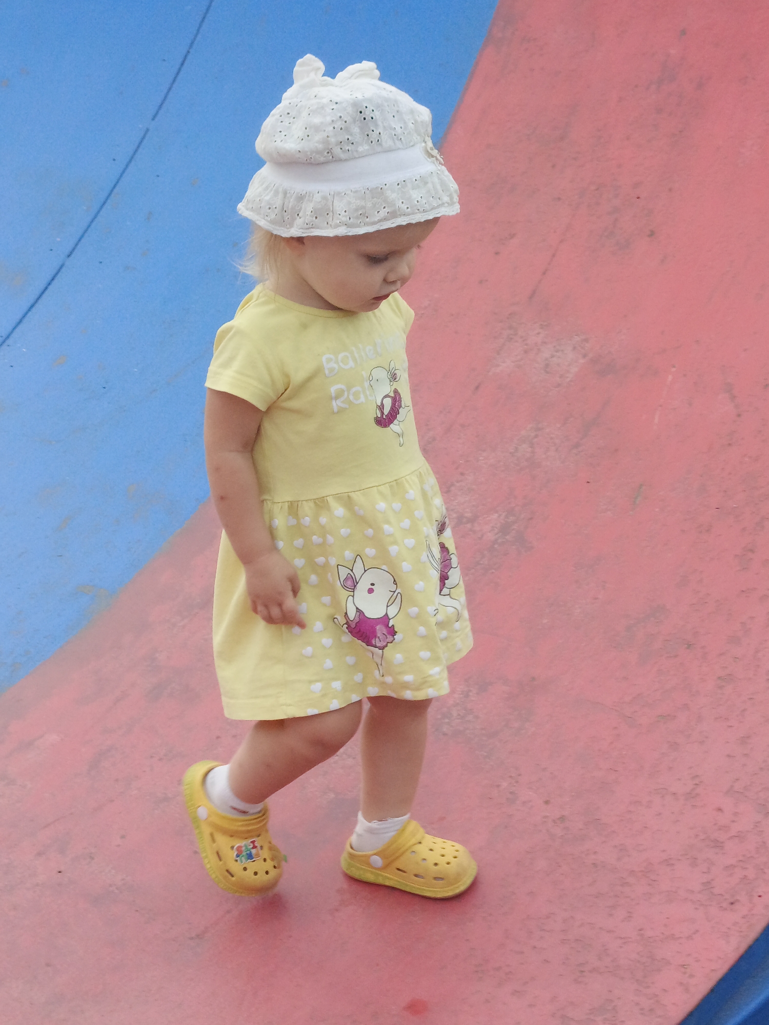 In Zlatoust, Russia, a young girl wearing a white hat, a yellow dress with a pattern of small white hearts and cartoon figures, and yellow Crocs, walks on a red and blue surface. The surface appears to be a slide or a playground structure. The girl is looking down and to her right, with her right leg extended forward.