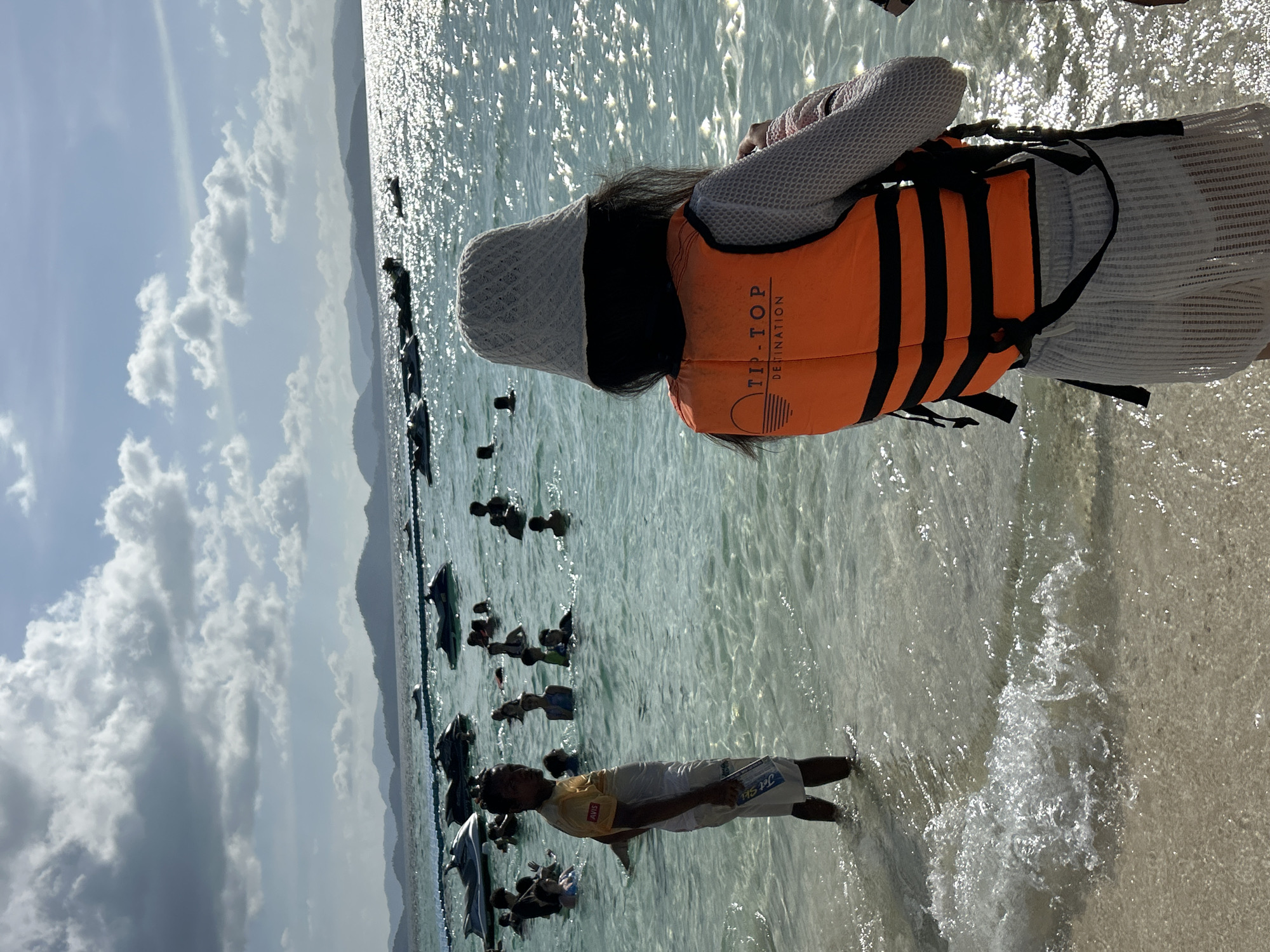 The image depicts a beach scene at Phru Nai, Thailand, with a clear, shallow sea and a sandy shoreline. In the foreground, a person wearing a white bucket hat, a light-colored long-sleeved shirt, and white textured shorts stands facing away from the camera, with an orange life vest over the shirt. Another person, a man, stands in the water to the left, wearing a yellow t-shirt with "AVIS" visible and white shorts. The sea is populated with numerous individuals, some swimming and others near a line of jet skis docked in the water. The background features a mountainous coastline under a sky with scattered clouds. The water is calm, with sunlight reflecting off its surface.