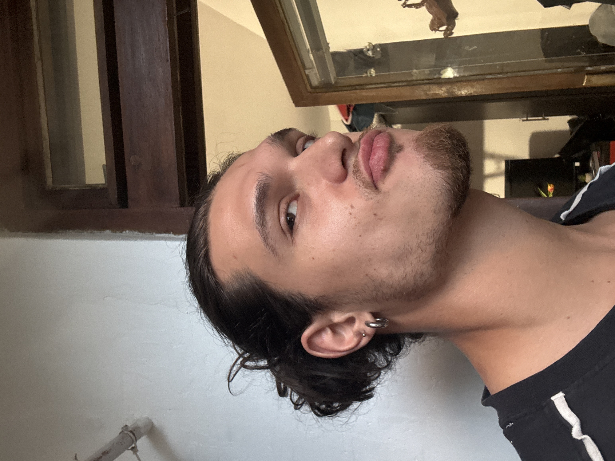 A man with dark, wavy hair, brown eyes, and stubble is looking up and to the left. He has earrings in his left ear. He is wearing a black shirt with a white stripe on the shoulder. The background includes a white wall, a brown window frame, and a mirrored cabinet or display case reflecting objects and light. There is no indication of location within the city of Sofia, Bulgaria, based on the image.