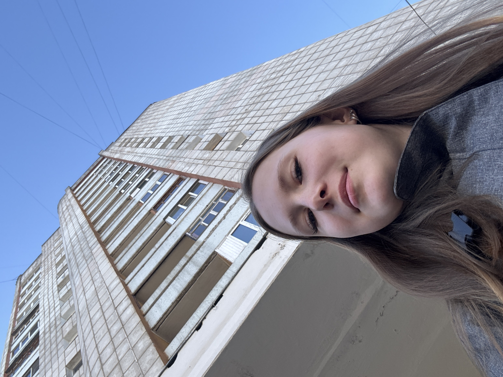 The image shows a young woman taking a selfie in front of a tall apartment building under a clear blue sky. The building is constructed from light-colored brick or concrete panels and has numerous windows. The woman has long brown hair and is wearing a grey jacket. The location is likely Chaikovsky, Russia, based on visual cues of architectural style, though this cannot be definitively confirmed. The angle of the shot suggests the picture was taken from a low vantage point, looking upwards.
