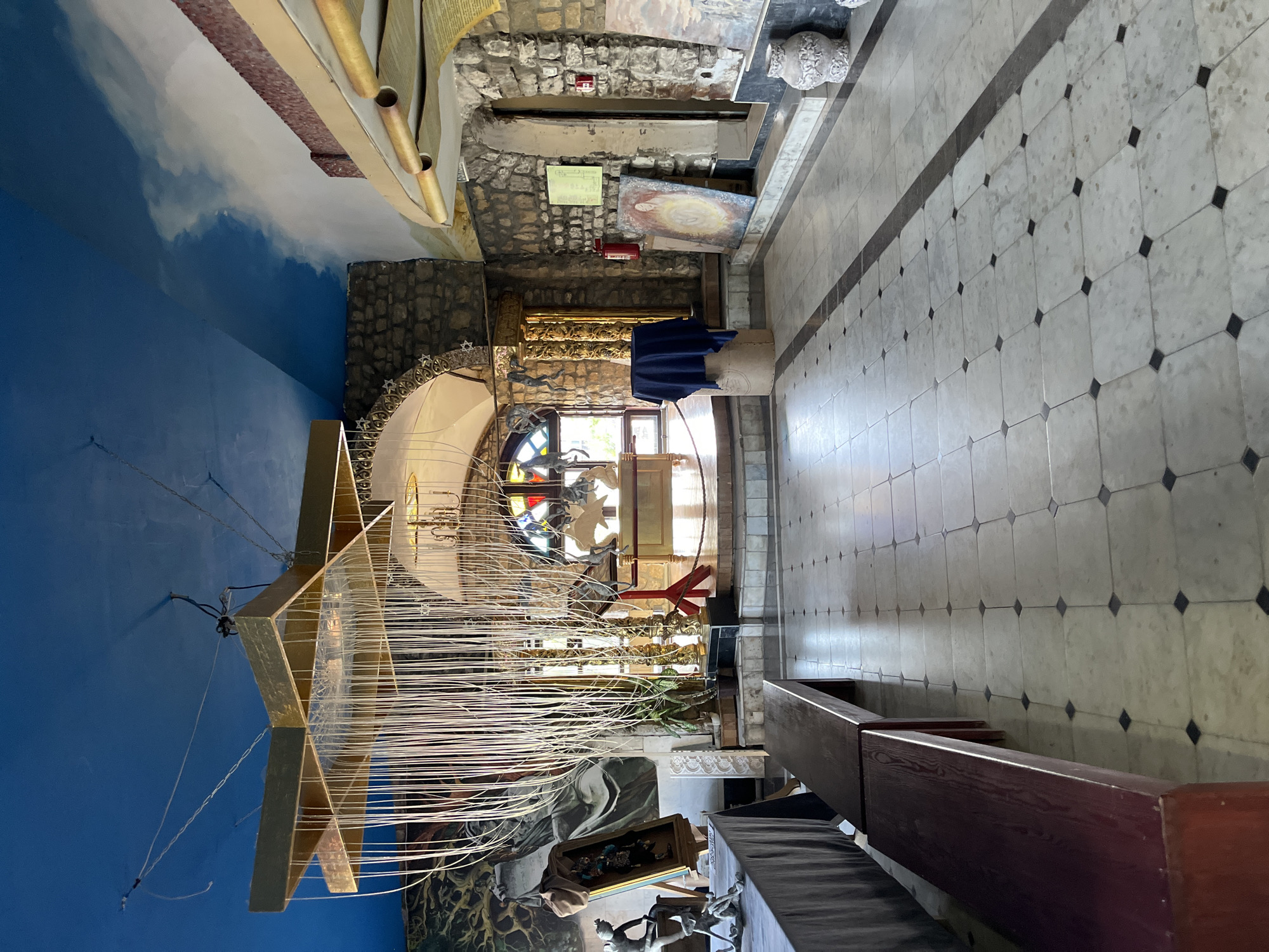 The image shows an interior space, likely a museum or art gallery, with a high ceiling painted with a sky motif. A large, star-shaped chandelier made of gold-colored material hangs from the ceiling, adorned with numerous white strands. The room is decorated with stone walls, arches, and various artworks, including paintings and sculptures. A long wooden bench is visible in the foreground, and a tiled floor with a diamond pattern covers the ground. In the background, a large arched window features stained glass. The overall impression is one of an eclectic and artistic environment. The location is identified as the Museum of Islamic Culture in Kazan, Russia.