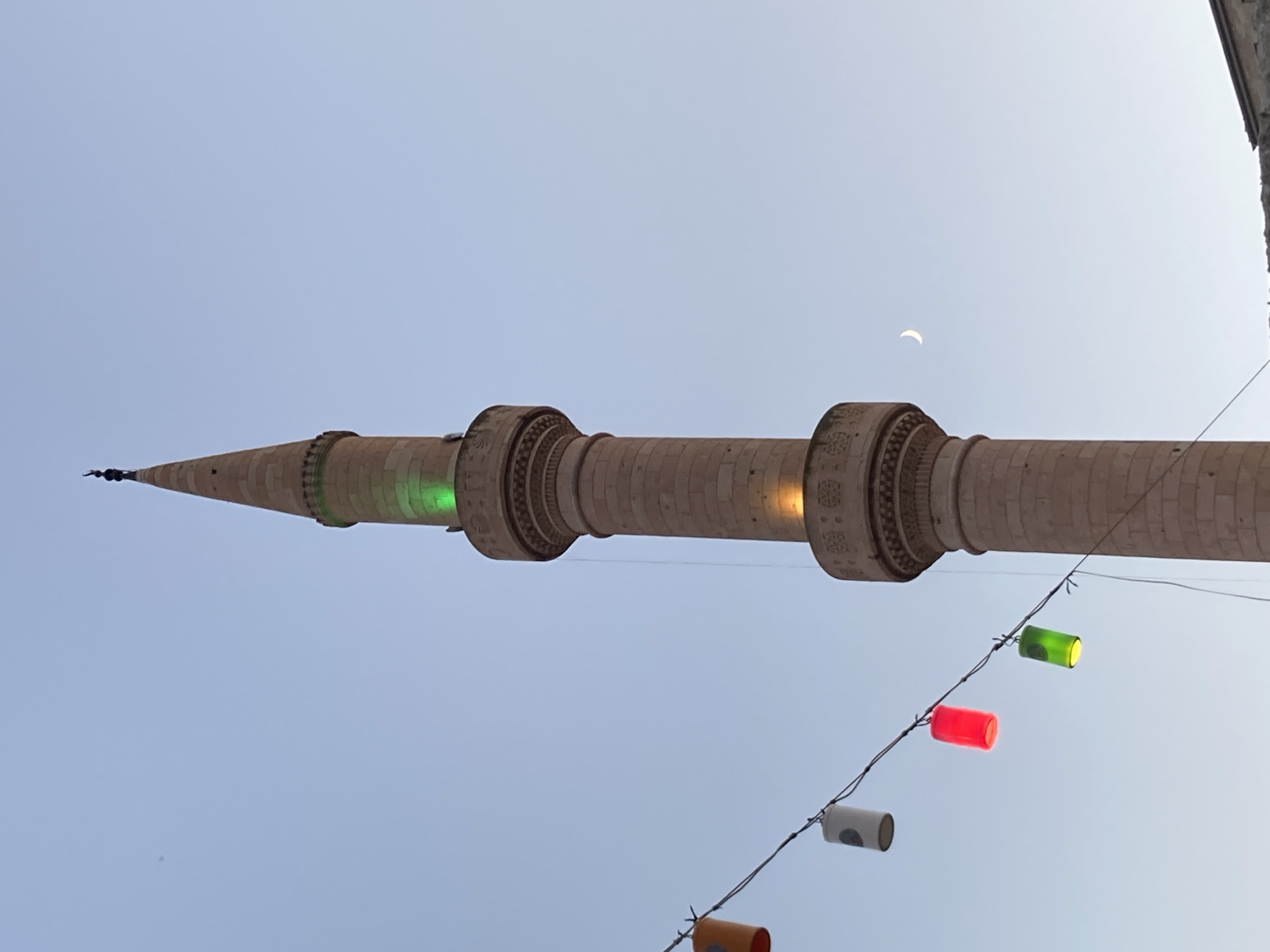 An image captured from below shows a minaret of a mosque against a clear, light blue sky. The minaret is constructed of light-colored stone and features intricate carvings at its base and mid-section. A string of colorful, cylindrical lanterns hangs from wires in the foreground, with red, green, and white lights visible. Green lights are also illuminating the upper portion of the minaret, and a single yellow light is visible lower down. A crescent moon is visible in the sky to the right of the minaret. The image appears to be taken in the city of Avanos İlçe Merkezi, Turkey. The bottom of the image shows a partial view of a building.