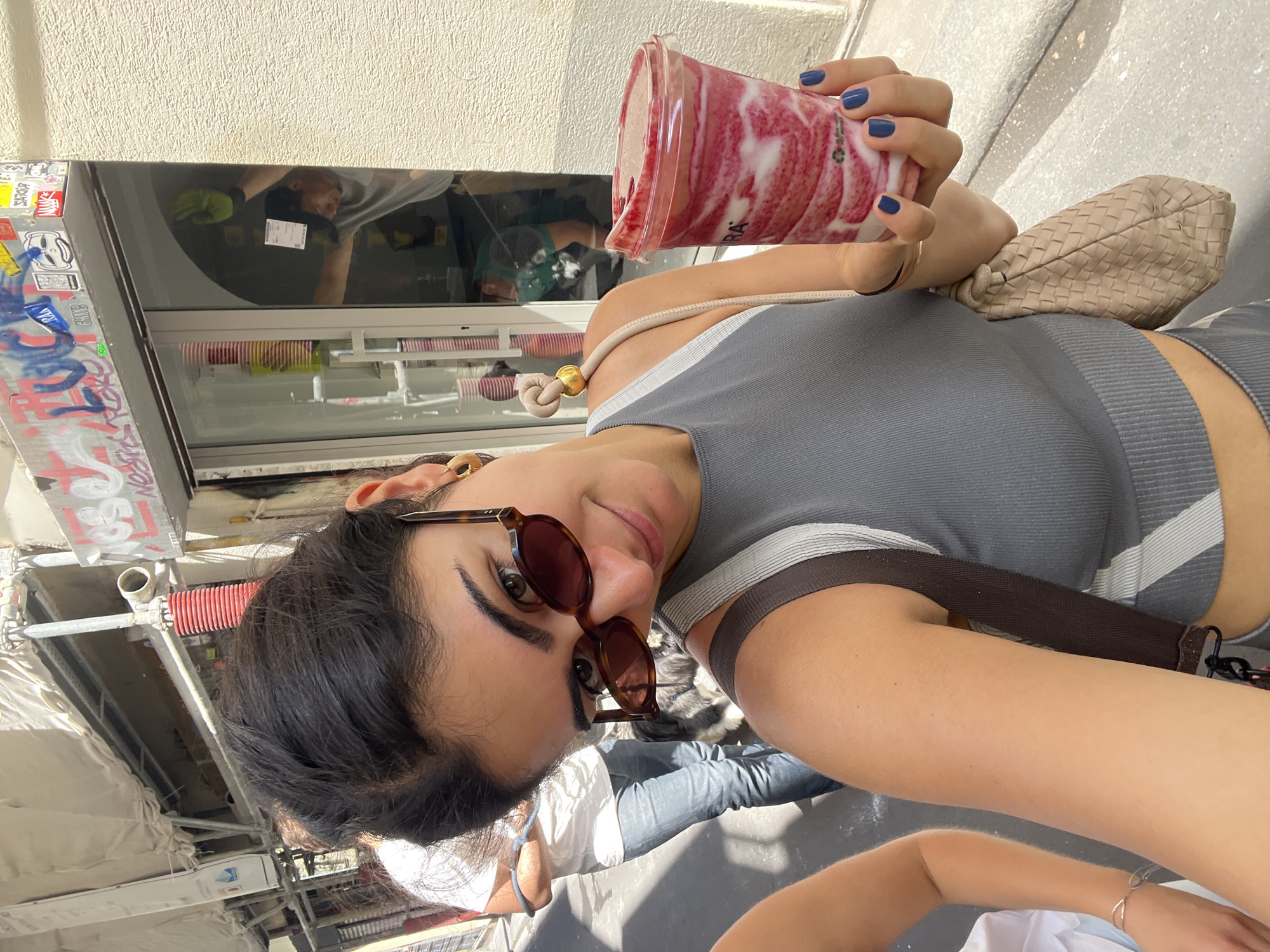 A woman with dark hair and sunglasses holds a pink and white swirl smoothie in a clear plastic cup in her right hand. She is wearing a gray tank top and a beige woven bag is on her left shoulder. She is standing on a street with a person wearing a white t-shirt and blue jeans walking behind her. In the background, there is a store with graffiti on the window and a person wearing a green glove inside. The scene appears to be an urban environment, possibly a street in Paris, France, given the context of the building's architecture and the presence of graffiti.