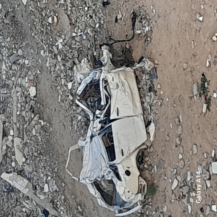 The image displays a severely damaged white four-door sedan situated on an expanse of uneven ground covered in dirt and extensive demolition debris in Khan Yunis, Palestinian Territory. The vehicle's roof is largely collapsed and heavily crushed inwards, with significant deformation visible across its entire body, including the front and rear sections. Twisted metal and exposed interior components are discernible. The surrounding environment consists of a densely scattered field of broken concrete fragments, rocks, and various unidentified rubble consistent with a destroyed area. Small patches of sparse vegetation are present on the dirt in the foreground. No individuals, actions, or interactions are observed in the static scene. A "Galaxy A14" watermark is visible in the lower-left corner of the image.