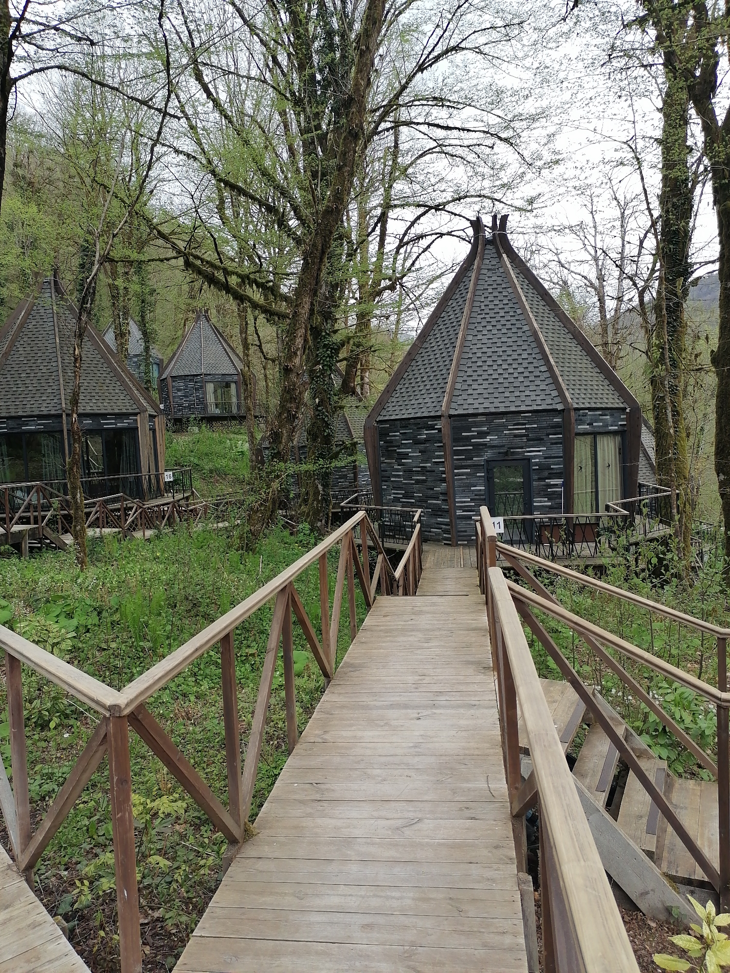 The image depicts a wooden boardwalk leading towards a cluster of small, uniquely designed cabin-like structures nestled within a forest environment. These cabins, identifiable by their dark, possibly shingled exteriors and distinctive pyramidal roofs, appear to be elevated, perhaps on stilts or constructed on a naturally sloping terrain. The boardwalk features wooden railings and appears to be descending towards the cabins. The area surrounding the boardwalk and cabins is densely wooded, suggesting a natural or semi-natural setting. The cabins appear to be situated in Medoveevka, Russia. The cabins have what looks to be room numbers in front of them. A number "11" is visible on the right side cabin. "16" is visible on the left side cabin. Other cabins can be seen on the background. The sky is overcast.