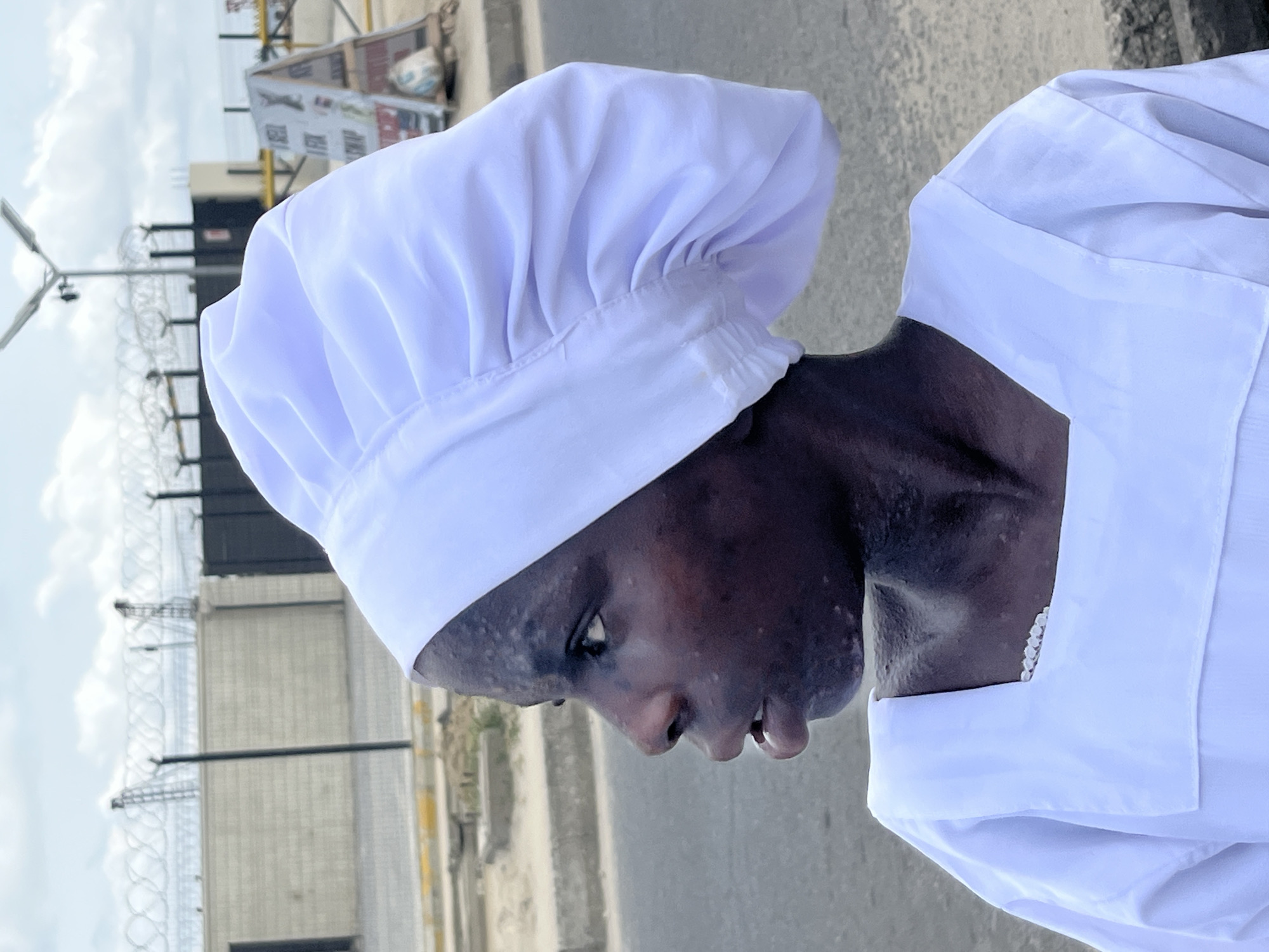 A person with dark skin is wearing a white chef's hat and a white uniform, viewed in a close-up, side-profile shot. The individual's gaze is directed downwards. The background is a blurred urban landscape featuring a street, barbed wire fencing, a light post, and a sign. The location appears to be Ado, Nigeria.