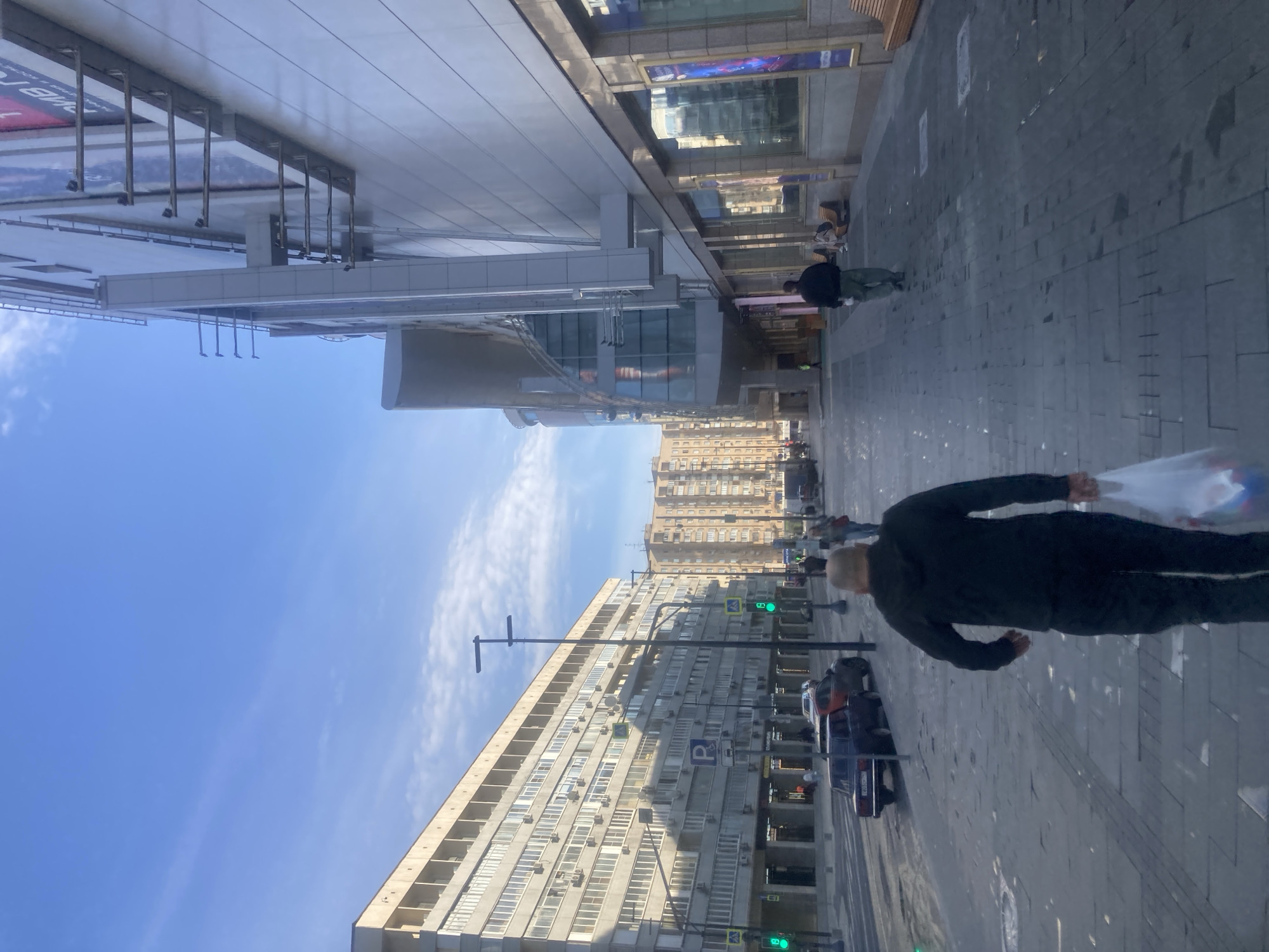 The image depicts a street scene in Moscow, Russia. The street is lined with tall, Soviet-era apartment buildings, characterized by repetitive window patterns and concrete construction. A modern shopping complex with a glass facade and external supports is visible on the upper side of the image. Pedestrians, including a man in dark clothing carrying a white bag, are walking on the paved sidewalk. Cars are parked along the curb. A street lamp and traffic lights displaying a green light are also visible. The sky is blue with scattered clouds.