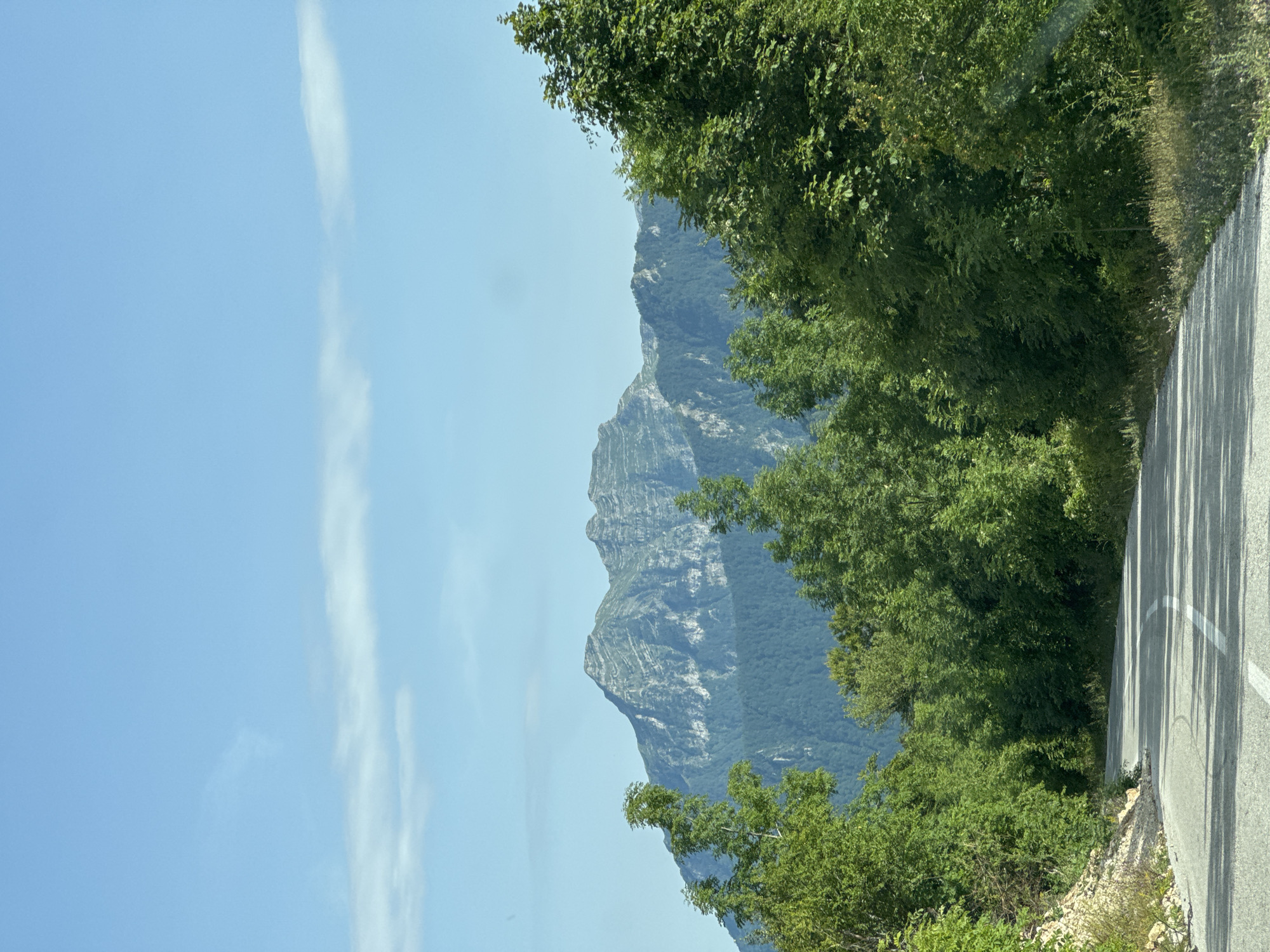 A paved road curves through a lush green forest, leading towards a majestic mountain range under a clear blue sky with scattered clouds. The road is marked with a white line down the center. The trees are dense and vibrant, with sunlight dappling through the leaves, casting shadows on the asphalt. In the distance, the rugged, layered slopes of the mountains are visible, their peaks softened by the haze. This scene is located in the vicinity of Dragovića Polje, Montenegro.