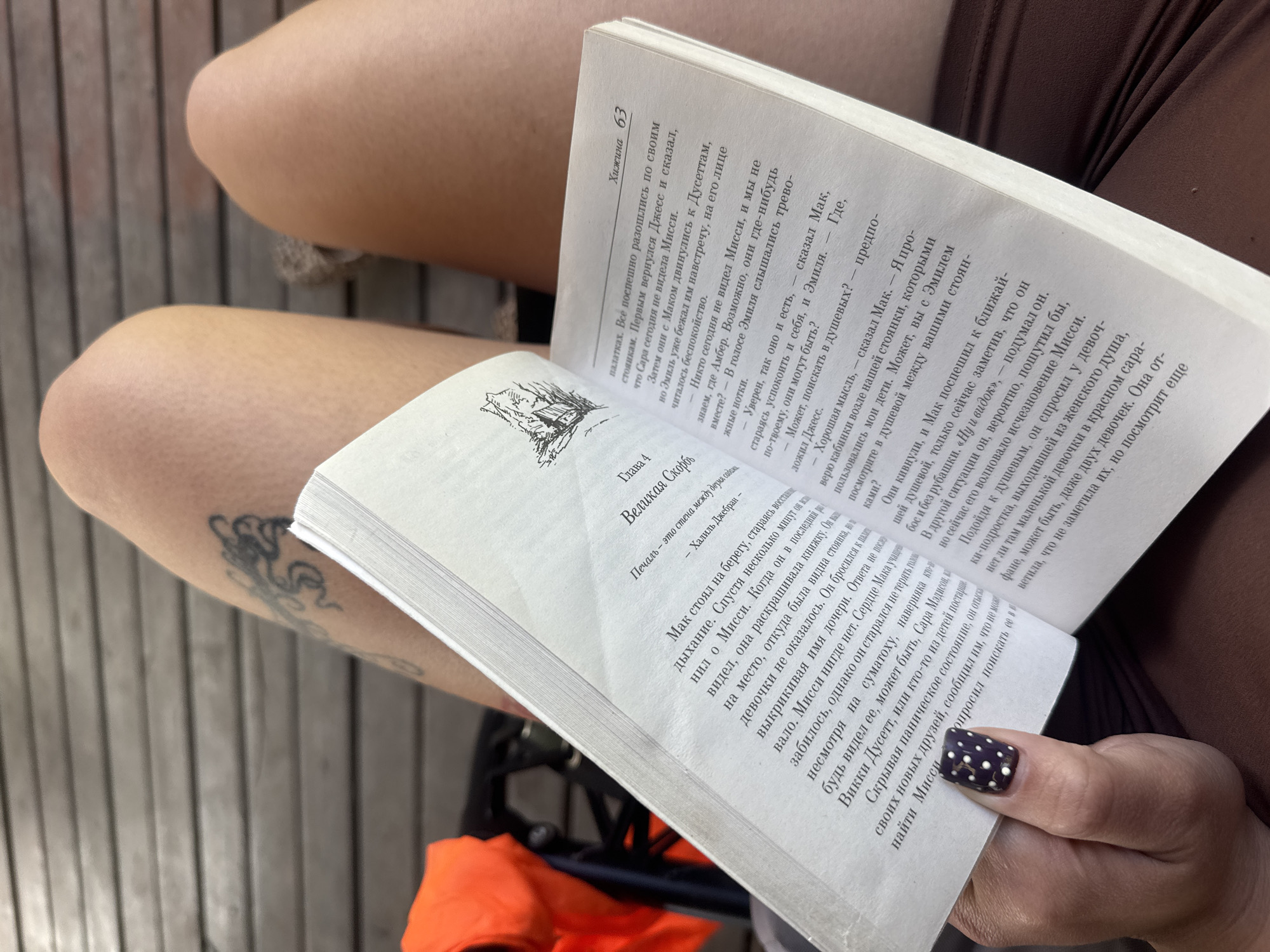 The image depicts a person, likely female, engaged in reading an open book. The person's bare legs are visible in the foreground, with an arm featuring a dark tattoo partially visible on the left side. The right hand, adorned with dark fingernail polish featuring white polka dots, gently holds the book open.
The book is a paperback with pages filled with Russian text. The right page is numbered '63' and has the word 'Хижина' (Hut/Cabin) at the top. The left page displays 'Глава 4 Великая Скорбь' (Chapter 4 The Great Sorrow/Grief) as a chapter title, beneath which is a quote: 'Печаль – это стена между двумя садами' (Sorrow is a wall between two gardens) attributed to Kahlil Gibran. A small line-drawing illustration, possibly of a cabin, is visible above the chapter title. The main text describes a narrative involving characters such as Missy, Mac, Jess, Emil, Amber, Sarah, and Vicky Doucett, focusing on a search for Missy and discussing checking showers, indicating a scene of concern or mild distress within the story.
The setting appears relaxed, possibly outdoors, with a background composed of horizontal wooden planks, suggesting a deck, floor, or wall. The lighting indicates daylight. Parts of brown and orange fabric are visible, likely belonging to the person's clothing or belongings. The overall impression is one of peaceful absorption in reading.