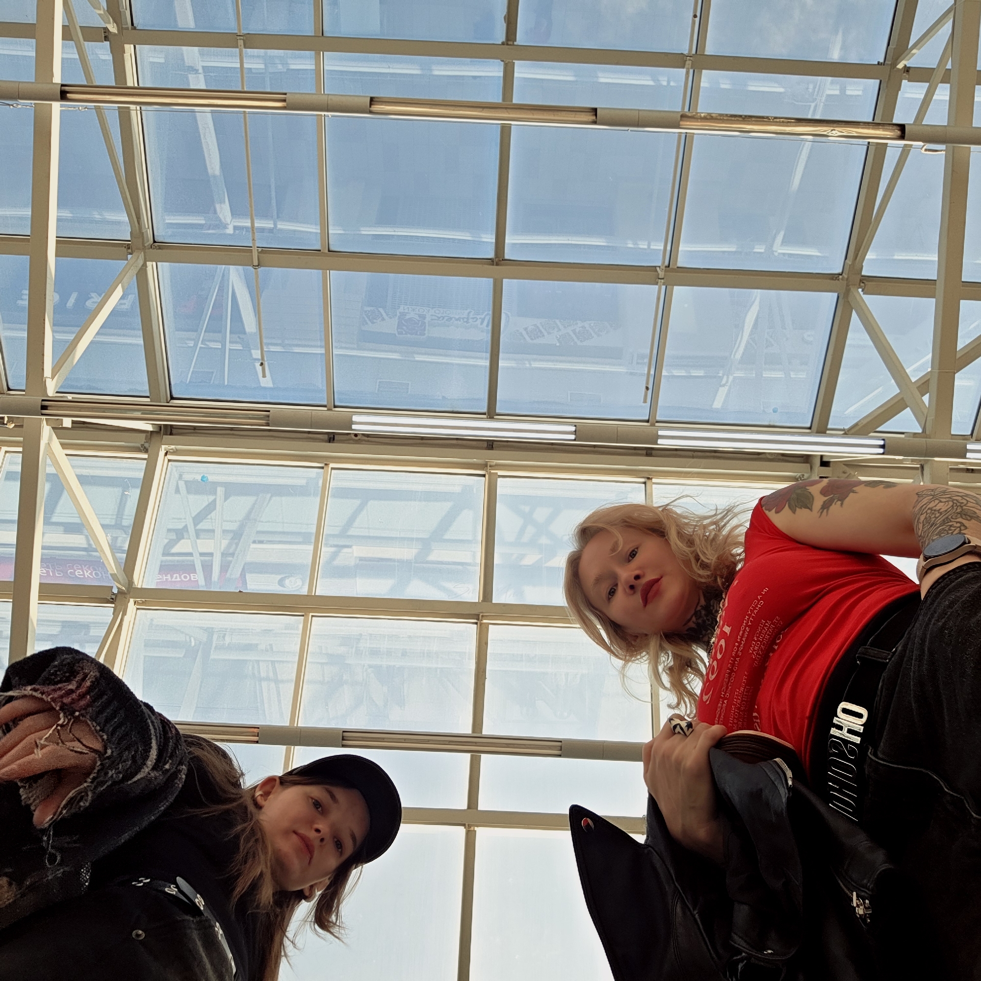 An upward-facing photograph captures two women looking down into the camera, likely in an indoor setting with a prominent glass roof, possibly a shopping mall or large modern building in Saint Petersburg, Russia. The bright, natural light filtering through the glass indicates it is daytime.
On the left, a woman with dark hair wears a black cap and dark clothing, possibly a jacket with distressed details on the sleeve. She looks directly at the camera with a neutral expression.
On the right, another woman with blonde, wavy hair wears a red t-shirt, black pants, and a black jacket draped over her arm. She also looks down with a serious expression, featuring red lipstick. Visible tattoos adorn both of her arms, one appearing to be a floral motif. A smartwatch is on her left wrist, and a silver belt buckle clearly spells out "MOSCHINO". The red t-shirt has white text printed on it, which is partially visible and mirrored, reading "A-2000 YOUTH AGAINST THE WORLD".
The background is dominated by the geometric pattern of the glass ceiling and its supporting metal framework. Reflections of signs and structures from outside or below are visible on the glass panels. Several mirrored texts can be deciphered, including what appears to be parts of Russian words like "брендов" (brands) and "есть секунд" (there are seconds), as well as "PRICE" and "МНОГО ЛОЖКИ" (many spoons). A rectangular lighting fixture runs across the ceiling structure.