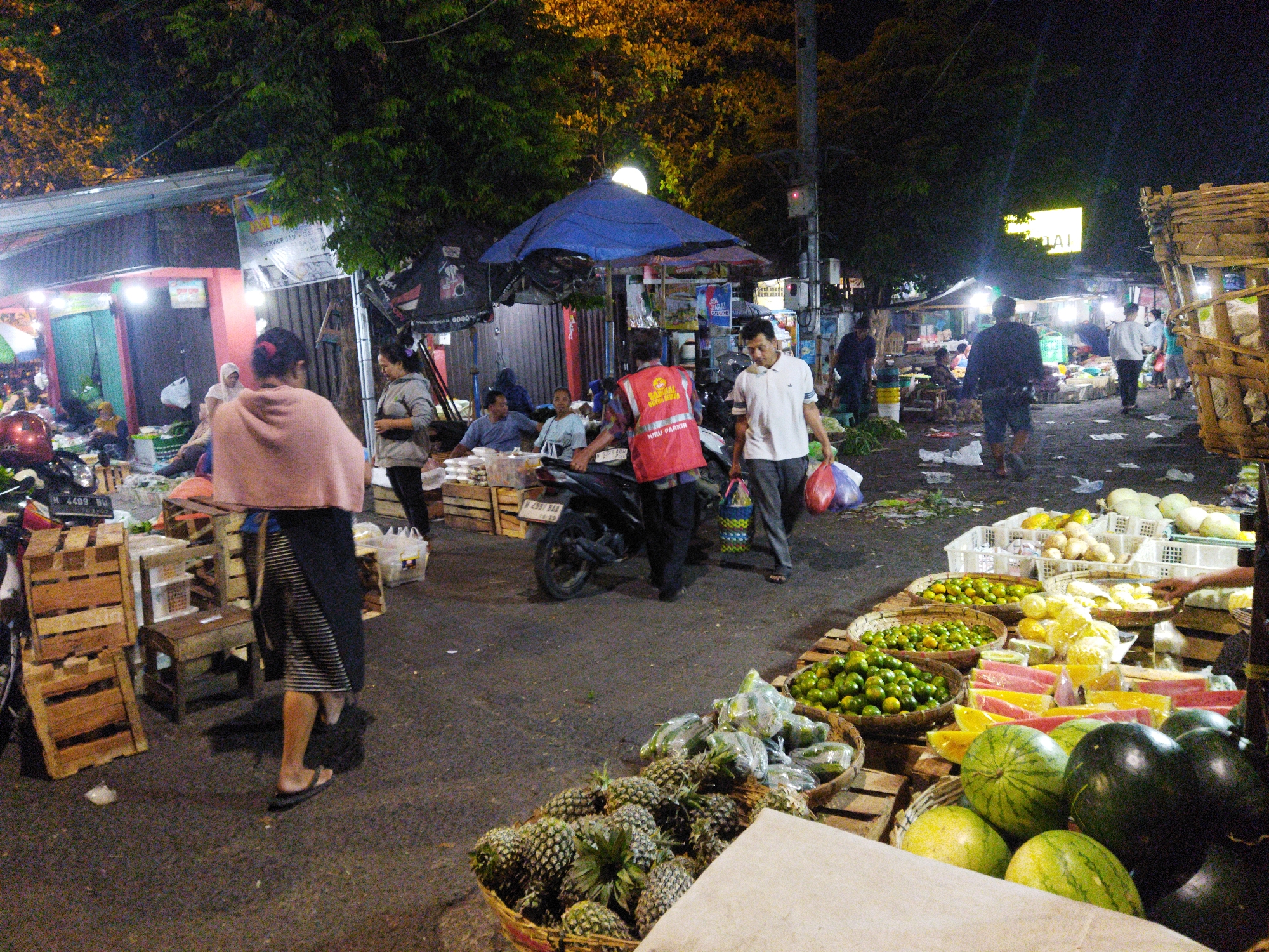 The image depicts a busy night market scene in Semarang, Indonesia. A variety of fruits are displayed in baskets and on wooden crates. In the foreground, there are large baskets of pineapples and watermelons. Further back, there are smaller baskets filled with oranges and other citrus fruits. Several people are seen browsing and shopping, while others are working at the stalls. One person is wearing a red vest with "POLISI" written on it, indicating a presence of law enforcement or market security. The market appears to be outdoors, with trees overhead and stalls made of wood and fabric. The lighting is dim, with some artificial lights illuminating the scene.
