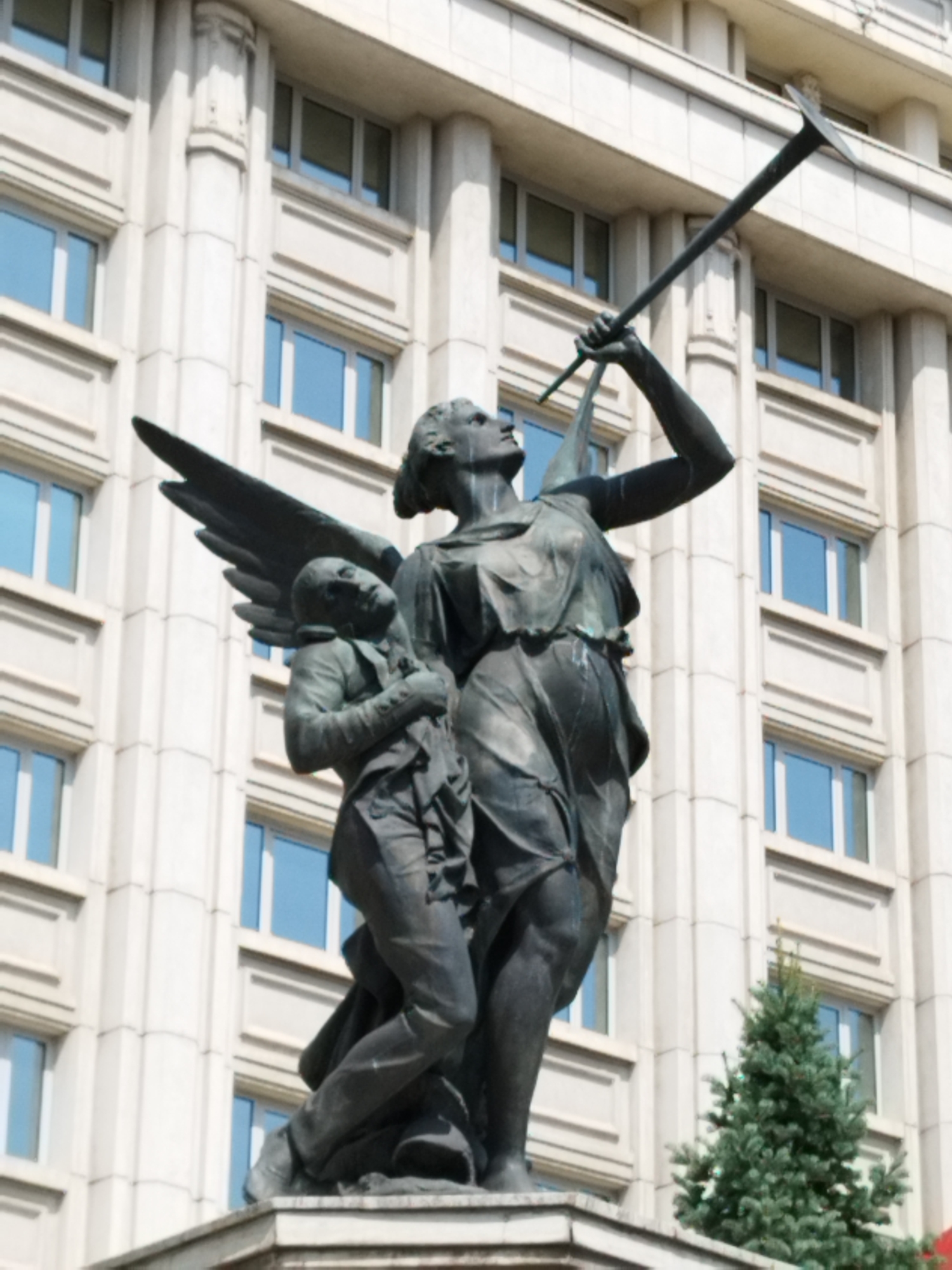 The image shows a statue of two figures in front of a building in Bucharest, Romania. The statue is made of dark metal and depicts an angel with wings, holding a long trumpet-like object. The angel is draped in flowing robes and is looking upwards. Beside the angel, a smaller figure, possibly a boy, is being held by the angel, with his head tilted back and his eyes closed. The building in the background is large and made of light-colored stone, with many windows. A small evergreen tree is visible to the right of the statue. The overall impression is one of solemnity and possibly reverence.